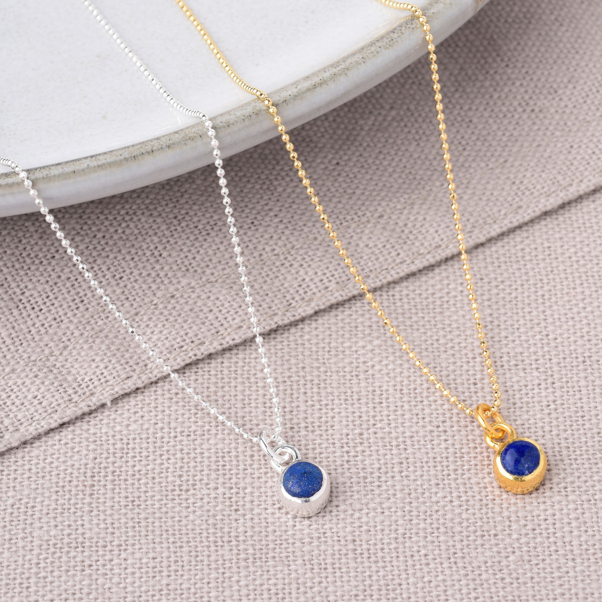 Two Gem Dot Birthstone Necklace September pieces, one in silver and one in gold, each featuring a small round blue lapis pendant, displayed on beige fabric with a white ceramic plate in the background.