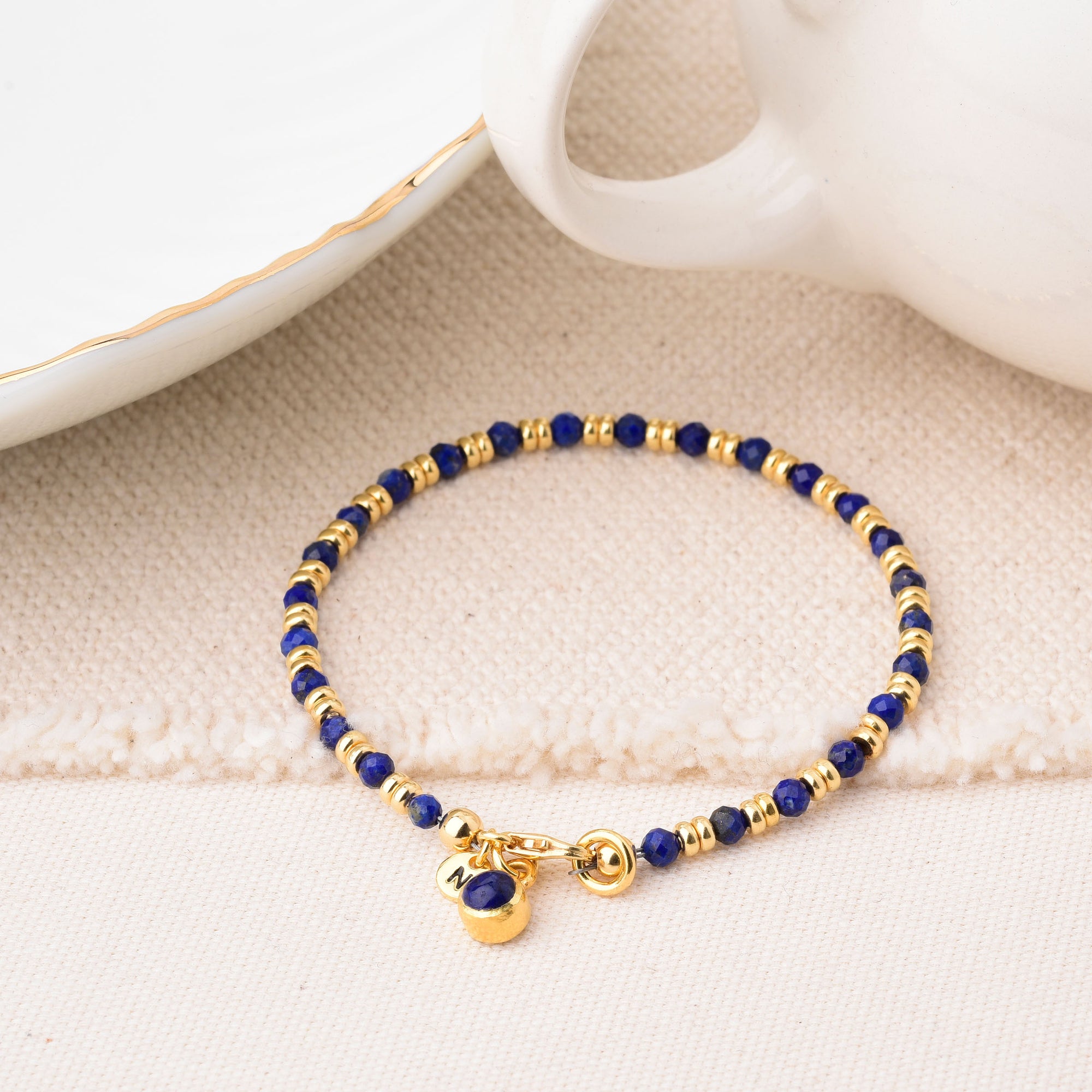 The Linear Birthstone Bracelet - September features alternating gold and dark blue lapis lazuli beads with a small gold charm, displayed on cream fabric beside a white plate and cup with gold trim.