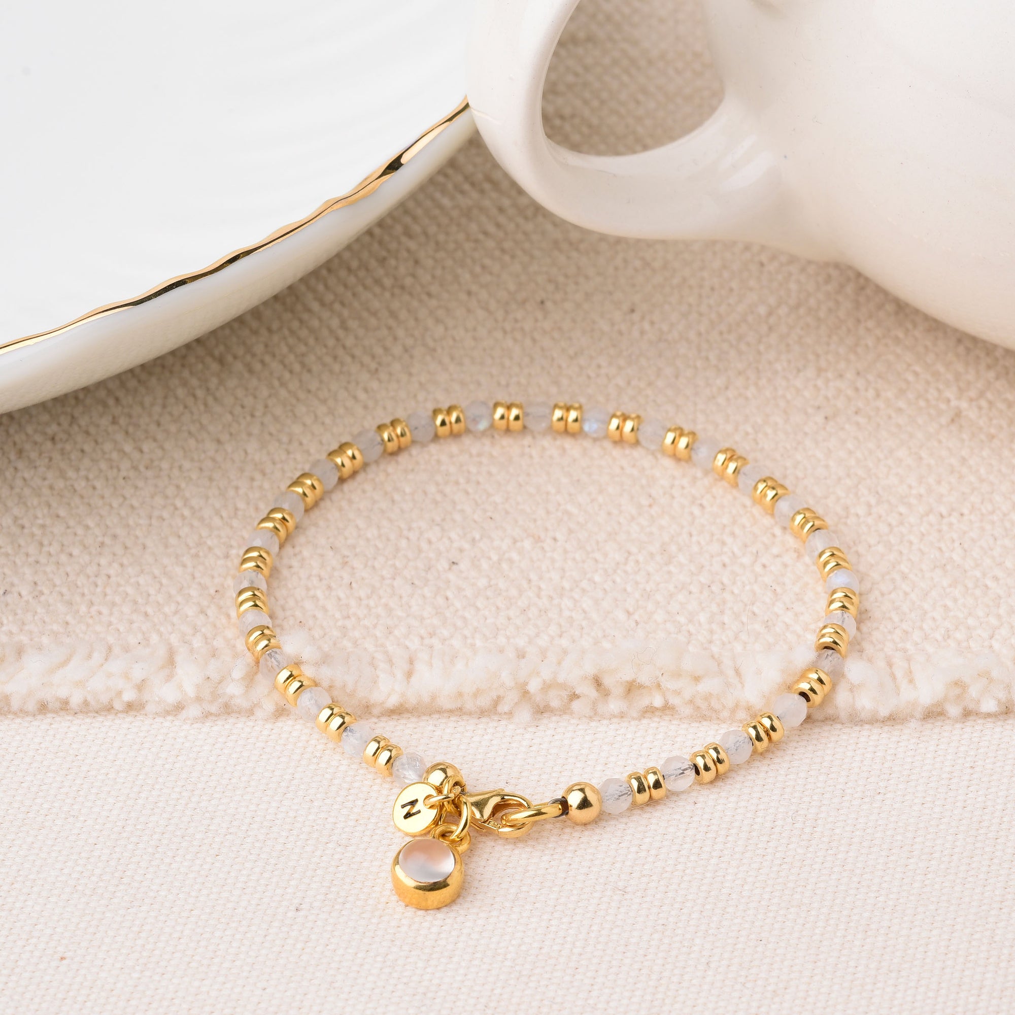 The Linear Birthstone Bracelet - June features gold and silver beads, a small pearl charm, and an engraved initial N charm. Shown on a cream textured surface next to white ceramic dishes with gold edges.