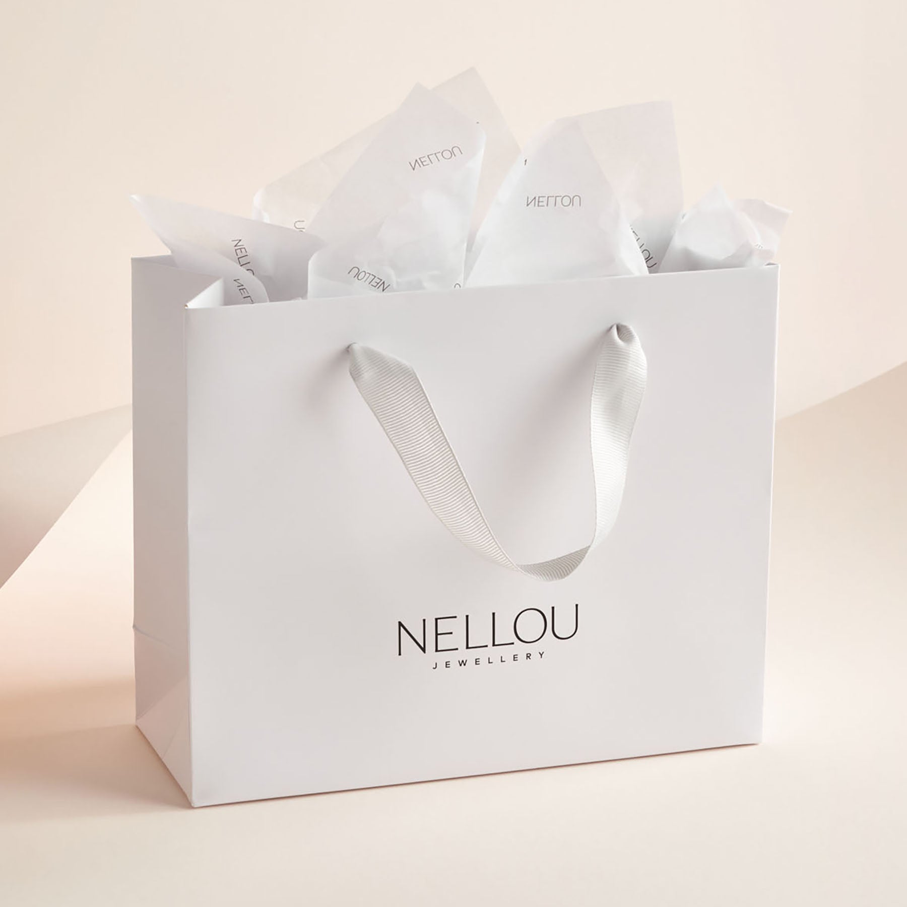 Luxury Gift Wrapping: A white gift bag with ribbon handles, filled with tissue paper and NELLOU JEWELLERY printed in black. The soft, neutral background highlights its elegant presentation.