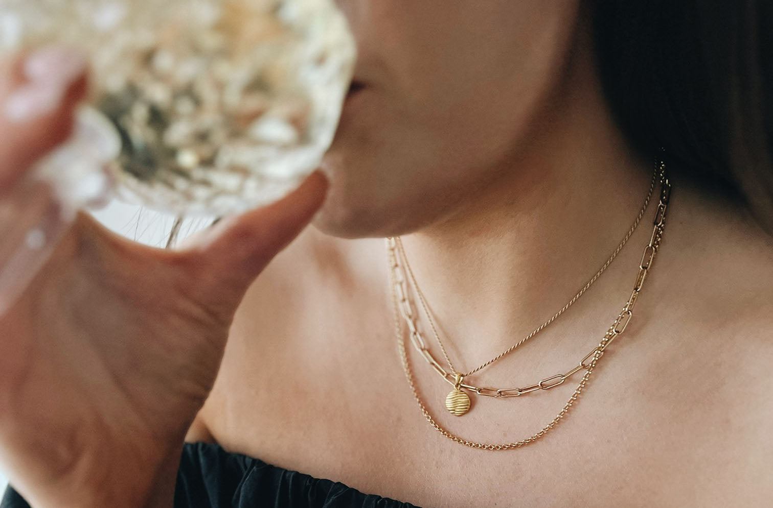 A woman wearing layered gold necklaces, one with a pendant, sips from a glass. She is dressed in an off-shoulder black top.