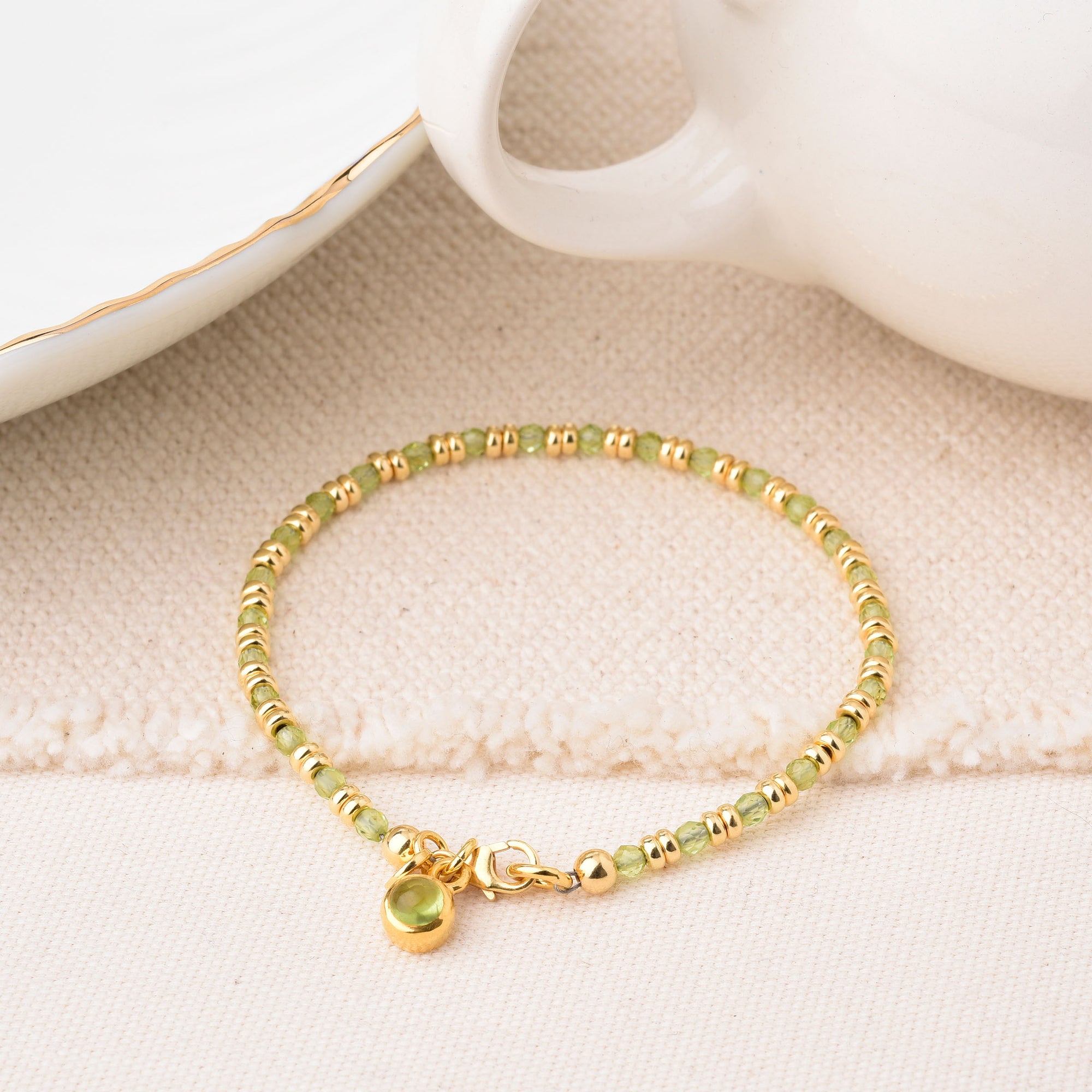 The Linear Birthstone Bracelet - August features alternating small gold and light green beads with a delicate gold charm, displayed on a cream textured surface beside a white teacup and saucer with gold trim.