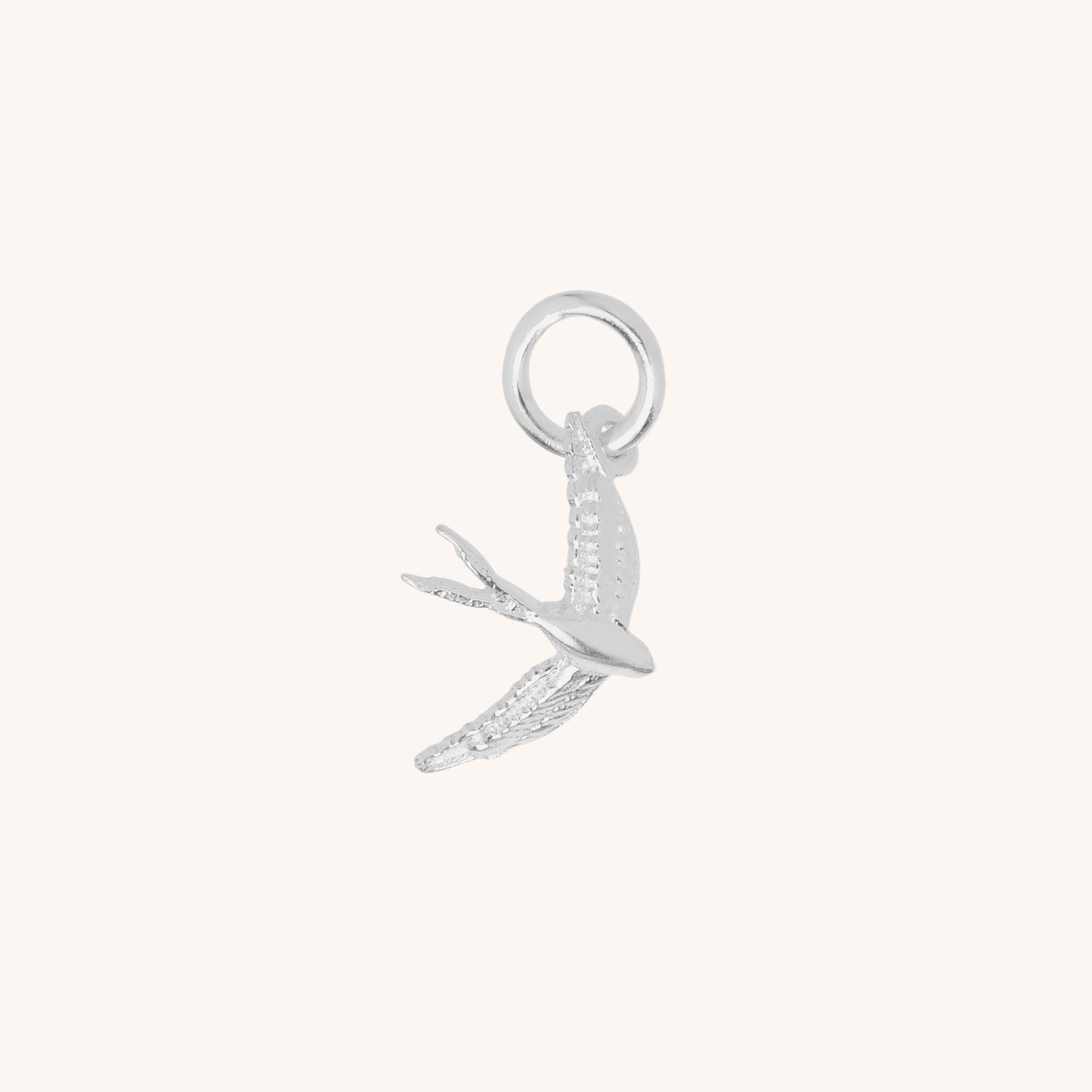 The Swallow Charm is a small sterling silver charm shaped like a flying bird with detailed wings, attached to a circular loop for hanging, shown on a plain light background.