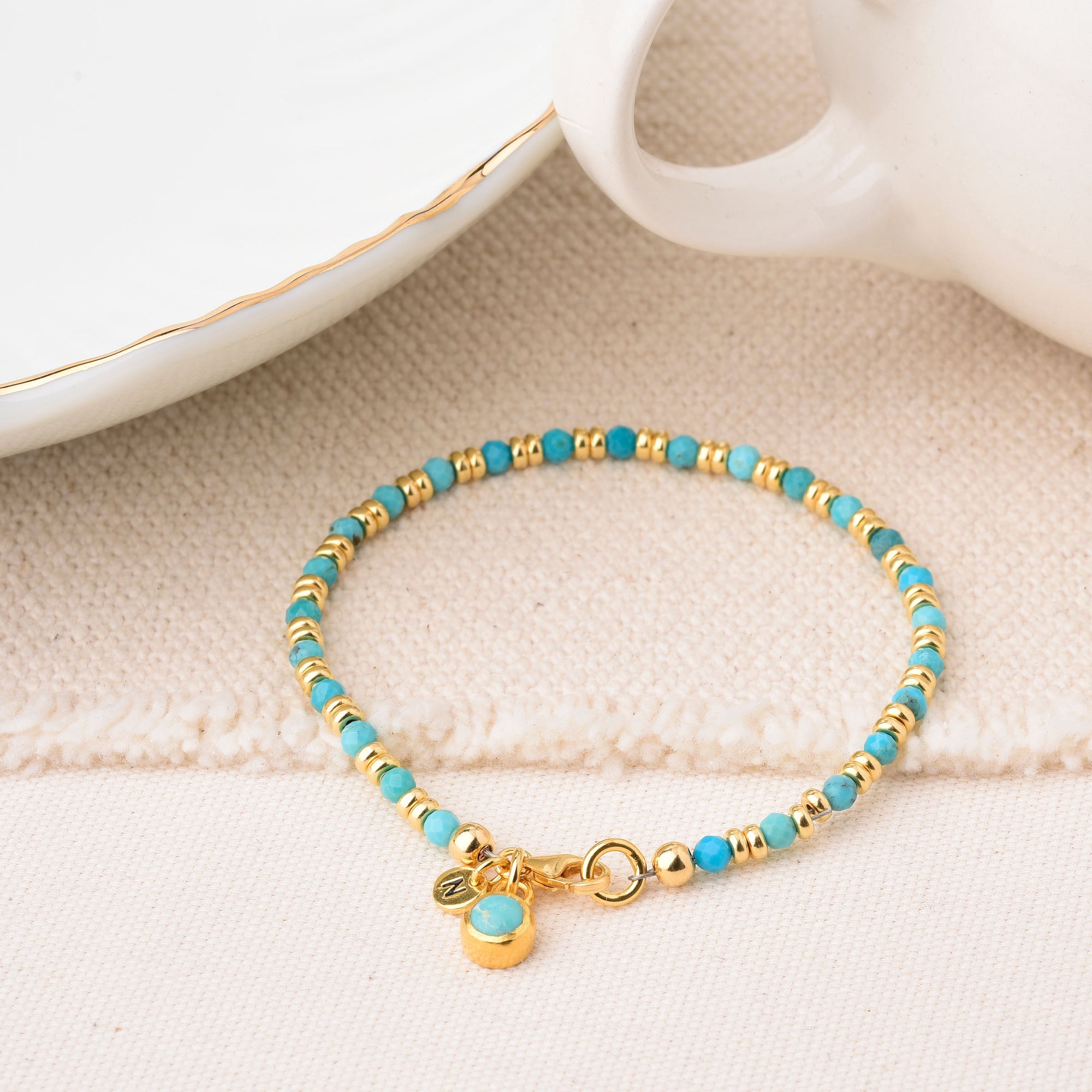 The Linear Birthstone Bracelet - December features turquoise and gold beads with a gold charm and turquoise pendant, displayed on textured fabric beside a white dish and gold-trimmed mug. Ideal for lovers of the December birthstone.