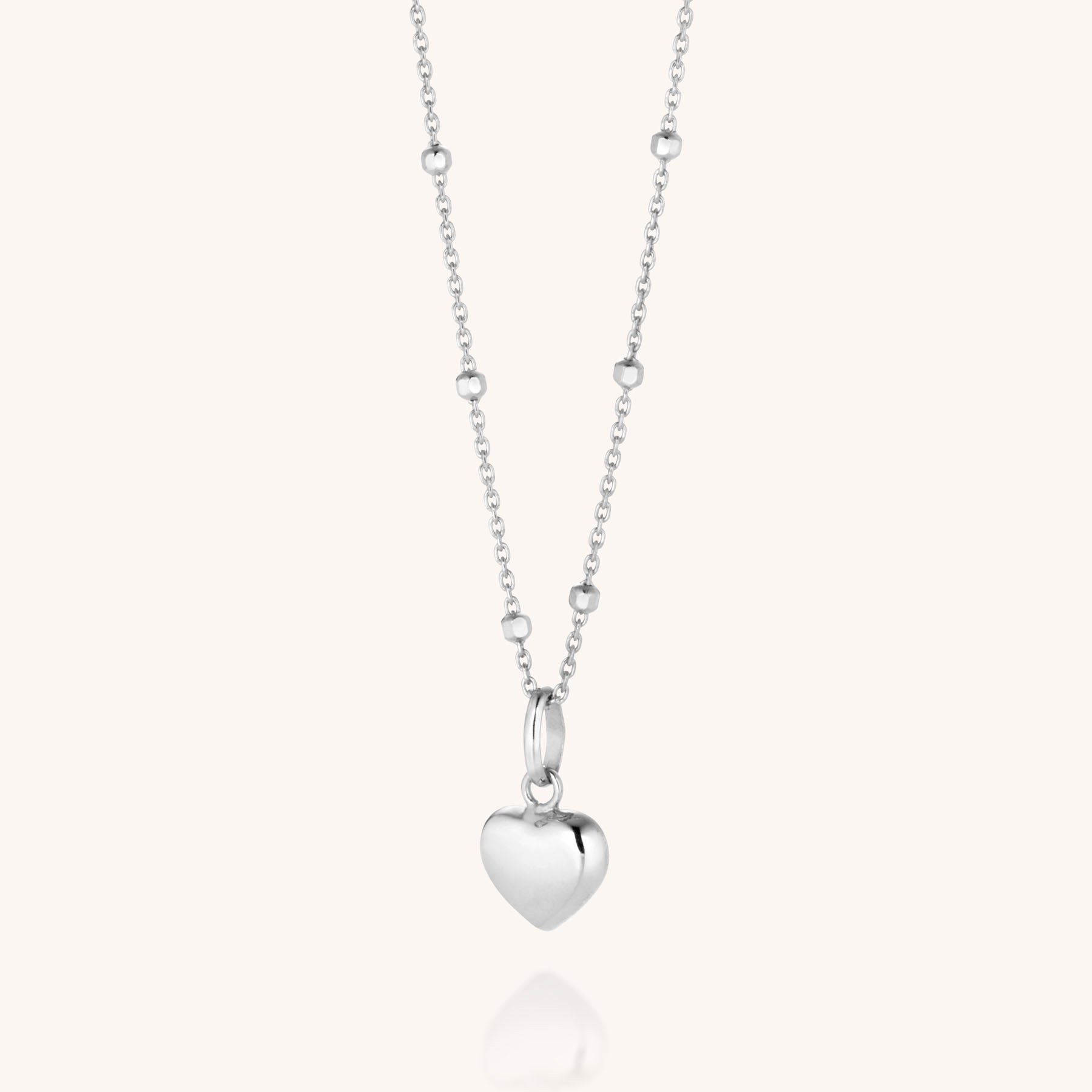 The Heart Charm Necklace is a delicate sterling silver piece with small beads along the chain and a polished heart pendant at the center, displayed on a soft off-white background.