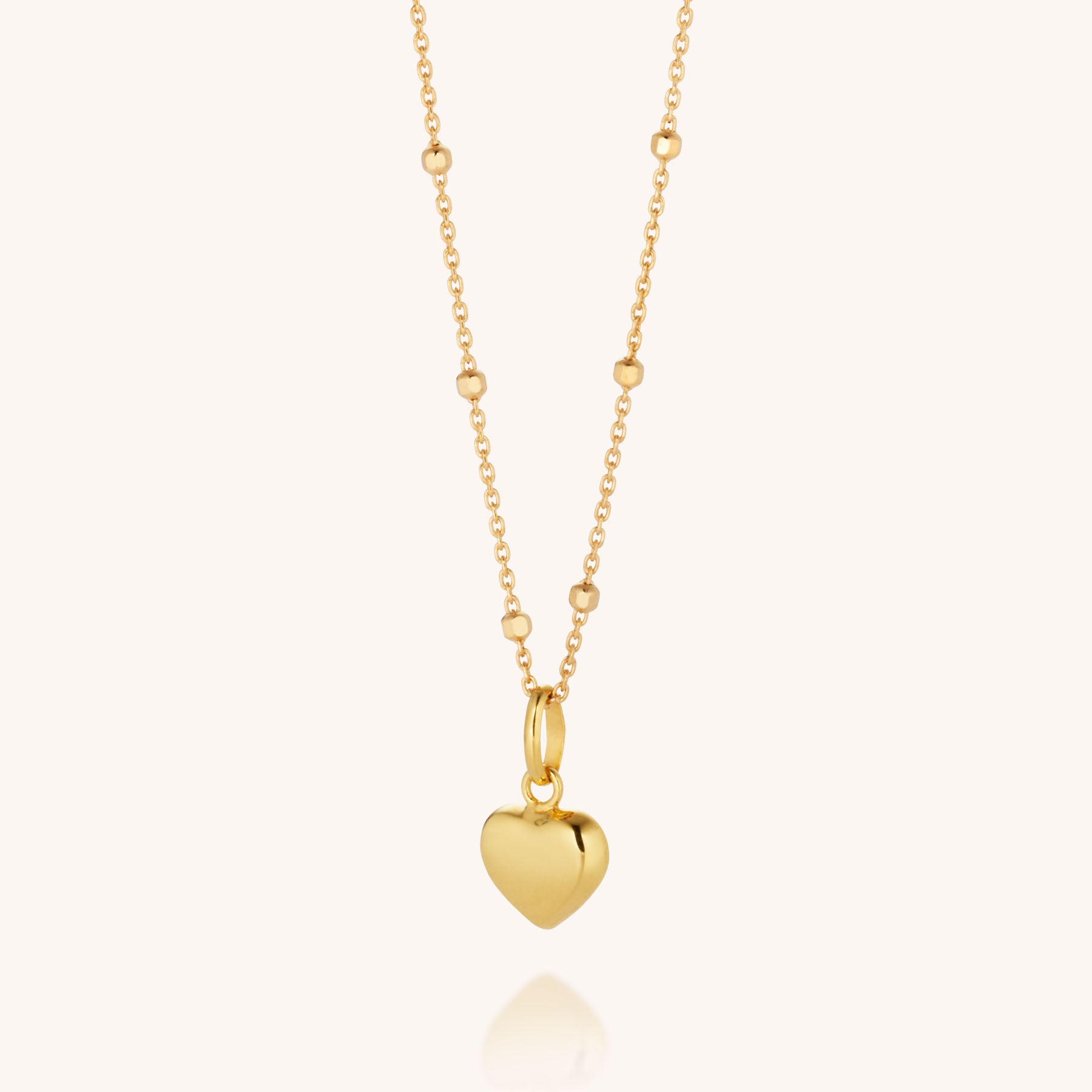 The Heart Charm Necklace is a gold plated beaded chain with a smooth heart pendant at the center, shown against a plain light background.