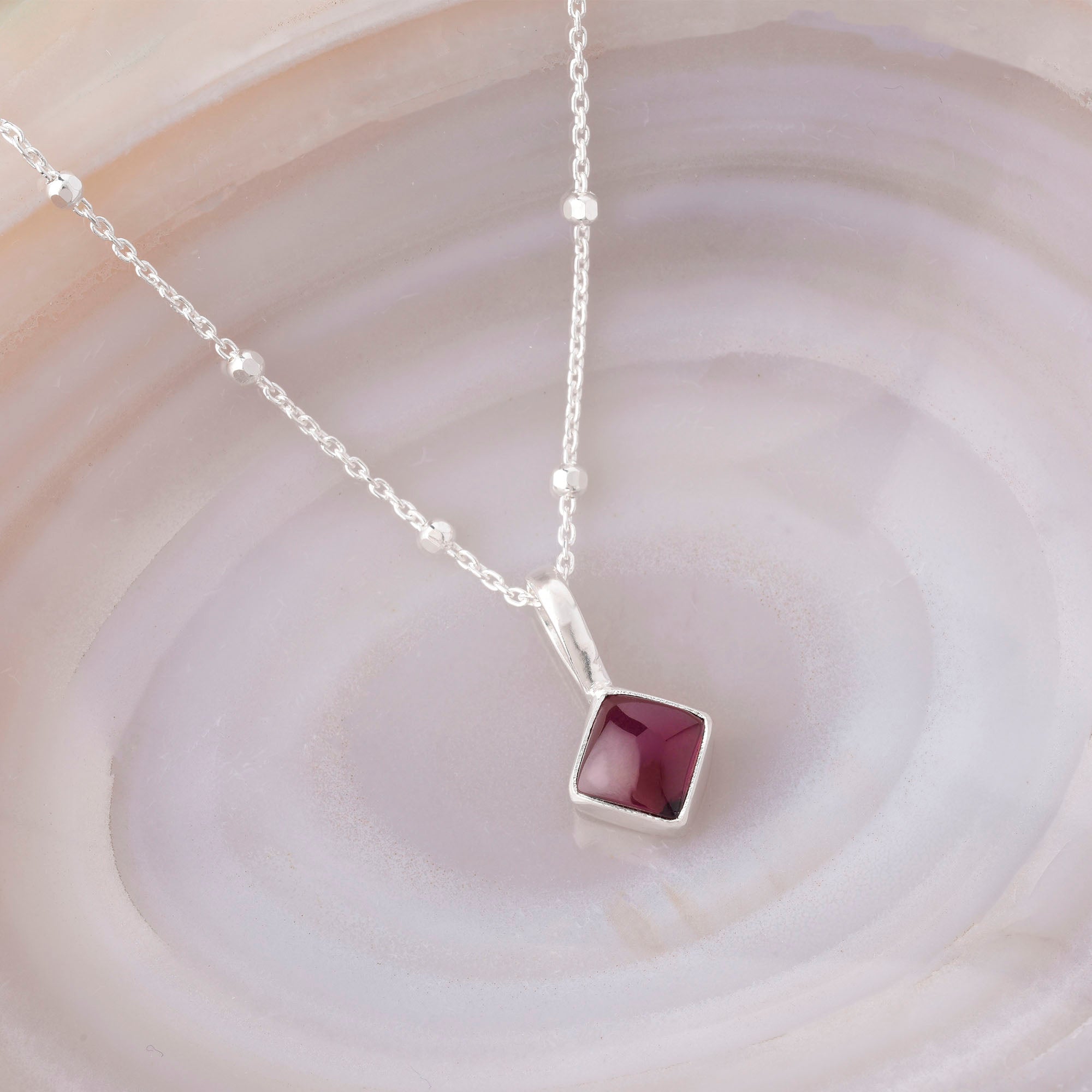 The Diamond Birthstone Necklace January is a delicate silver necklace with small beads and a square pendant set with a deep red Garnet, making it a thoughtful personalized birthday gift, displayed on swirled light-colored marble.