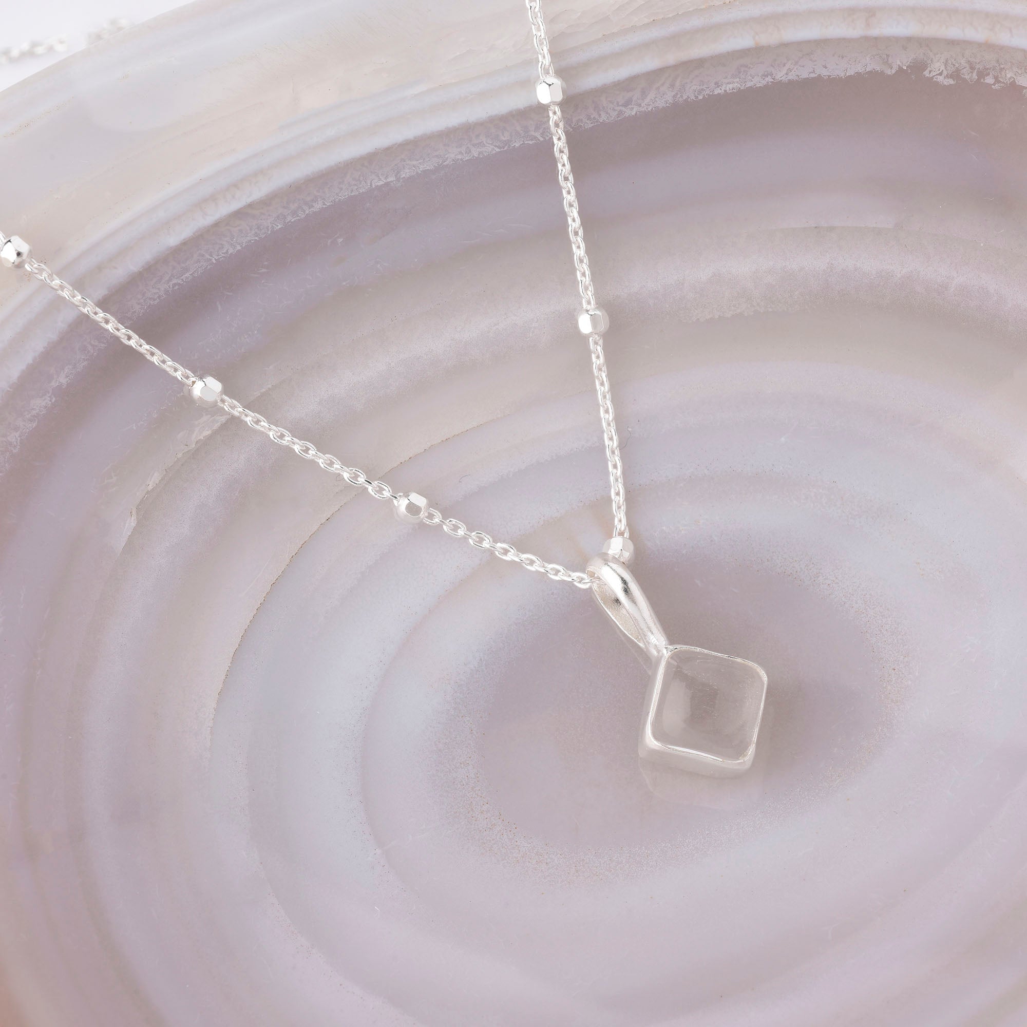The Diamond April Birthstone Necklace, a delicate silver piece with a small square pendant, makes a perfect personalised birthday gift, beautifully displayed on a soft gray and lavender banded agate stone background.
