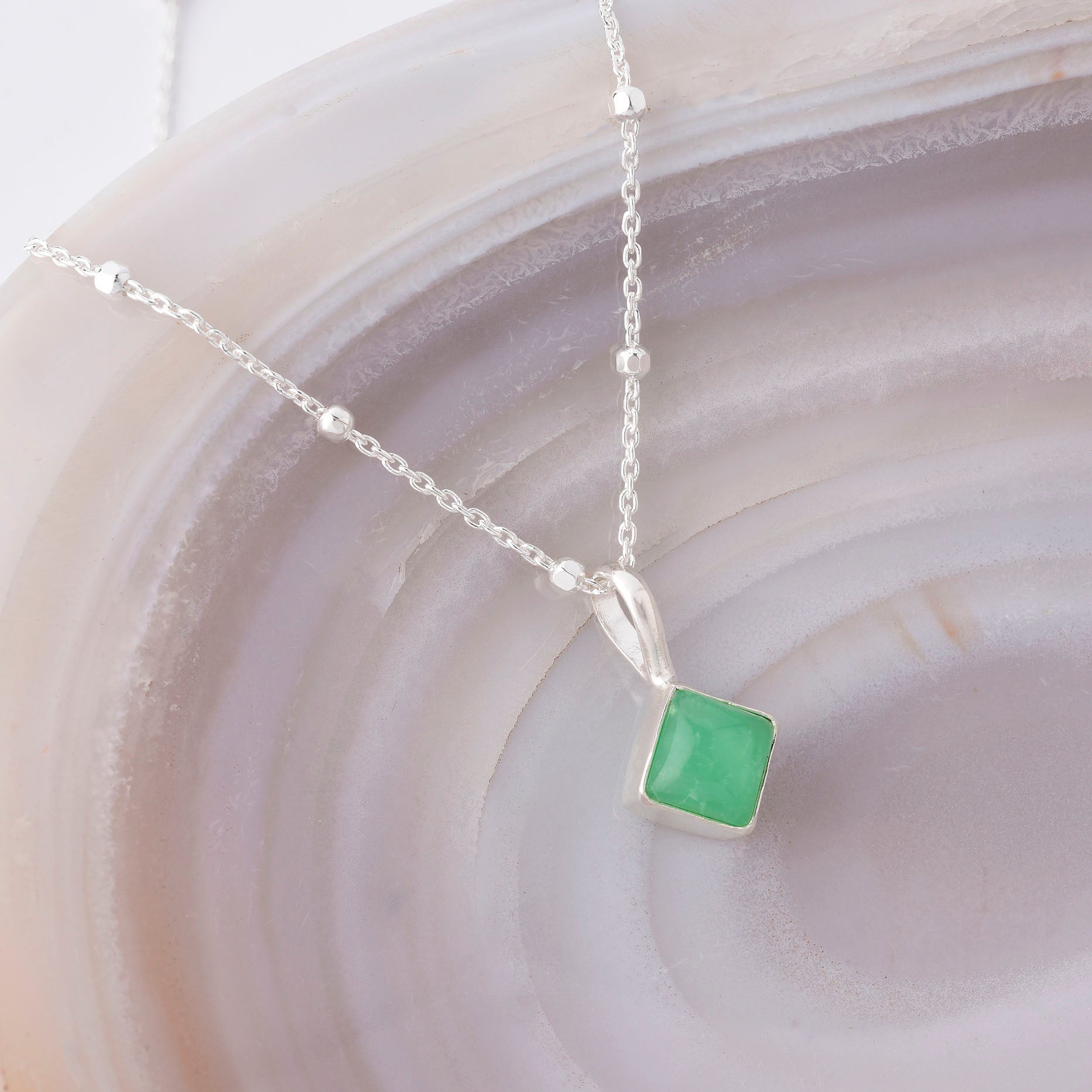 The Diamond Birthstone Necklace May, featuring a silver pendant with a square Chrysoprase green gemstone, is displayed on a polished white and beige stone surface, highlighting its unique beauty and healing properties.