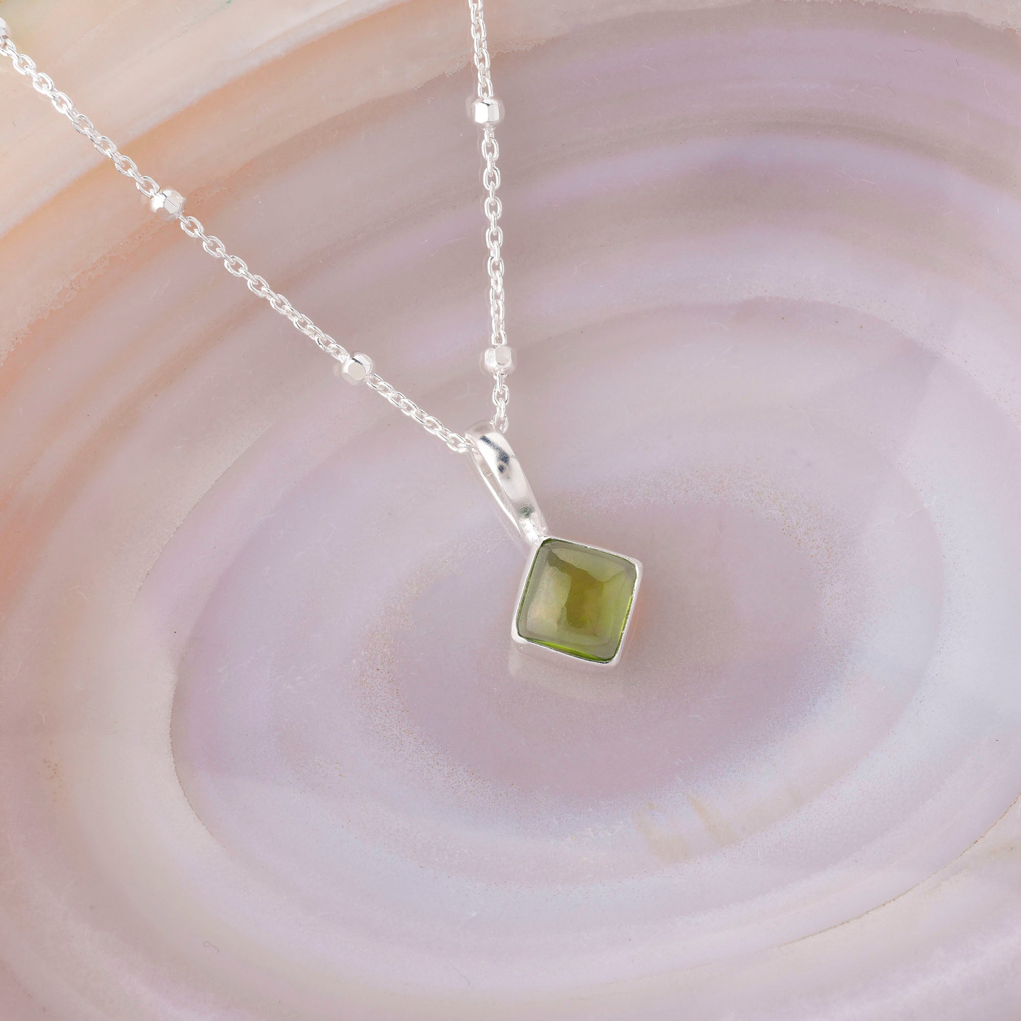 The Diamond Birthstone Necklace August, featuring a shimmering green gemstone pendant, is showcased on a soft pink and purple agate background, enhancing its elegance and the unique qualities of the August birthstone.