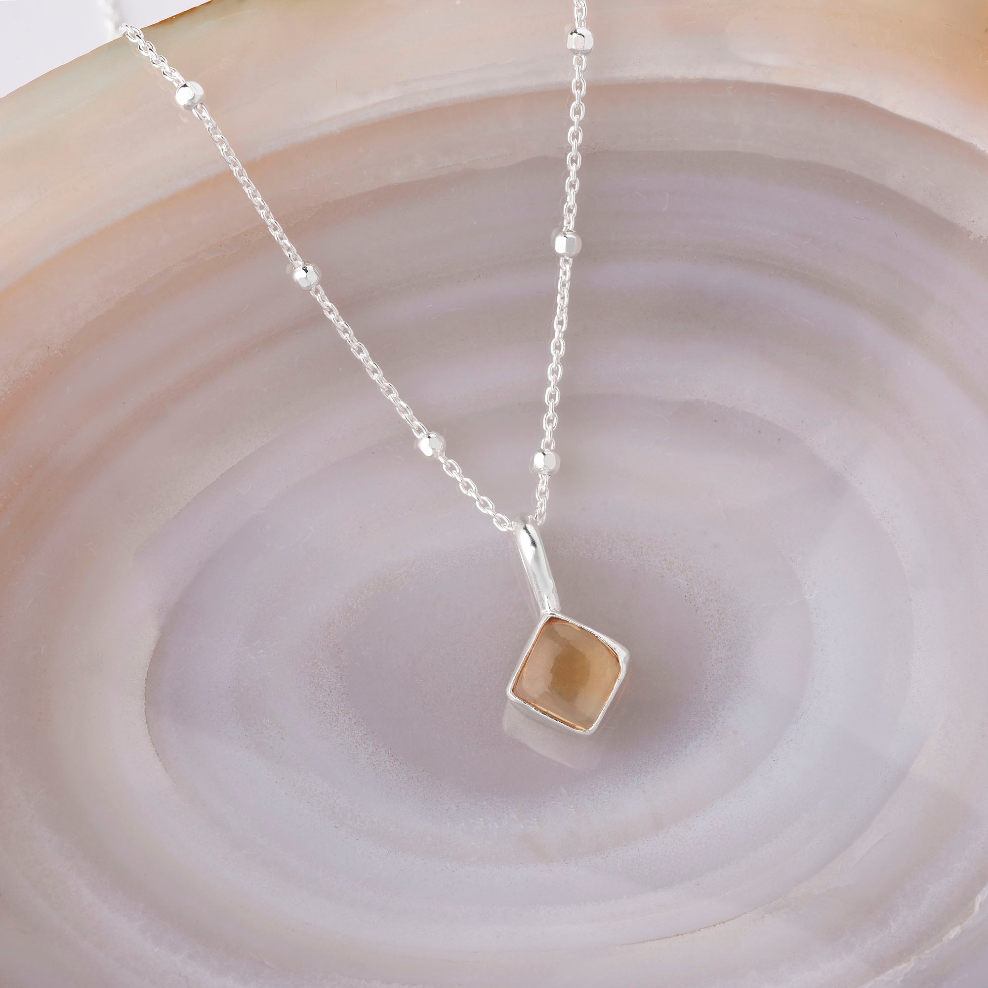 The Diamond Birthstone Necklace November is a delicate silver chain with small beads and a square pendant featuring a translucent brown birthstone, elegantly displayed on a smooth marble surface with beige and taupe swirls.