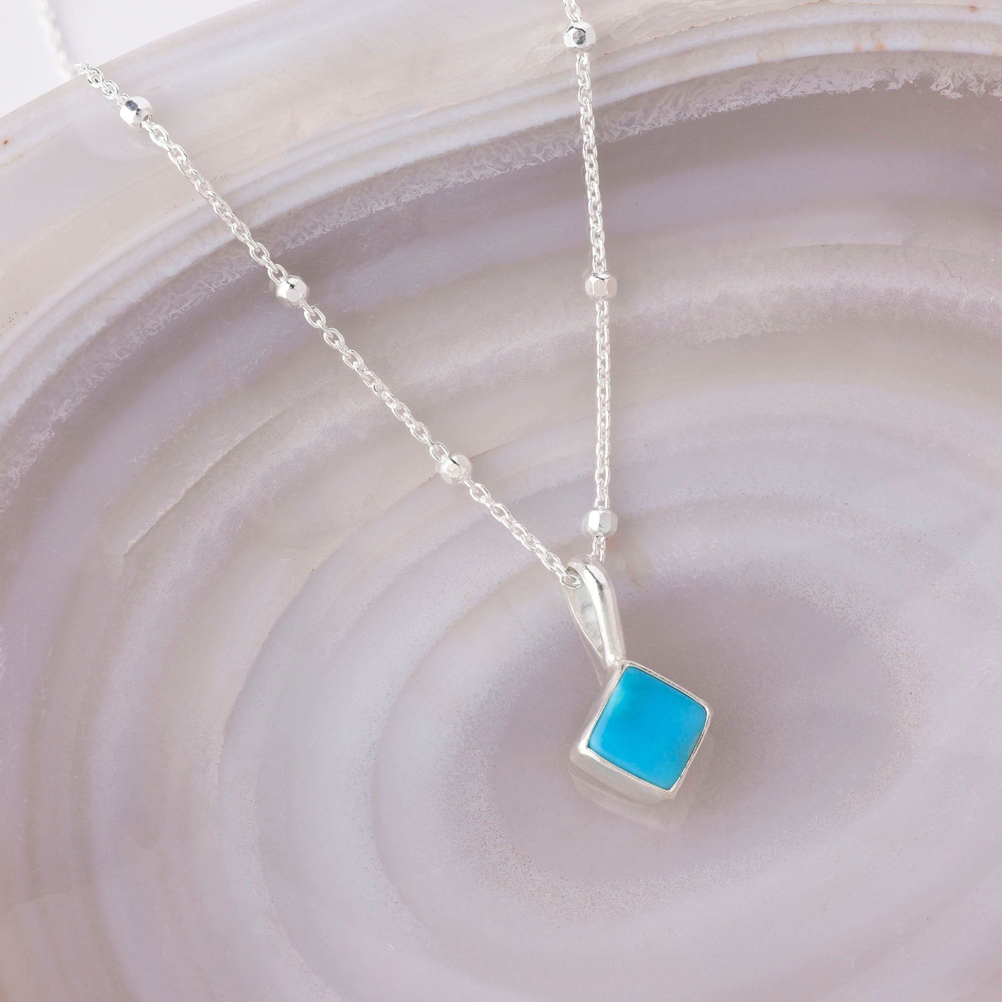 A delicate silver chain displays the Diamond Birthstone Necklace December, featuring a sparkling square pendant, beautifully showcased on a light-colored, banded stone background.