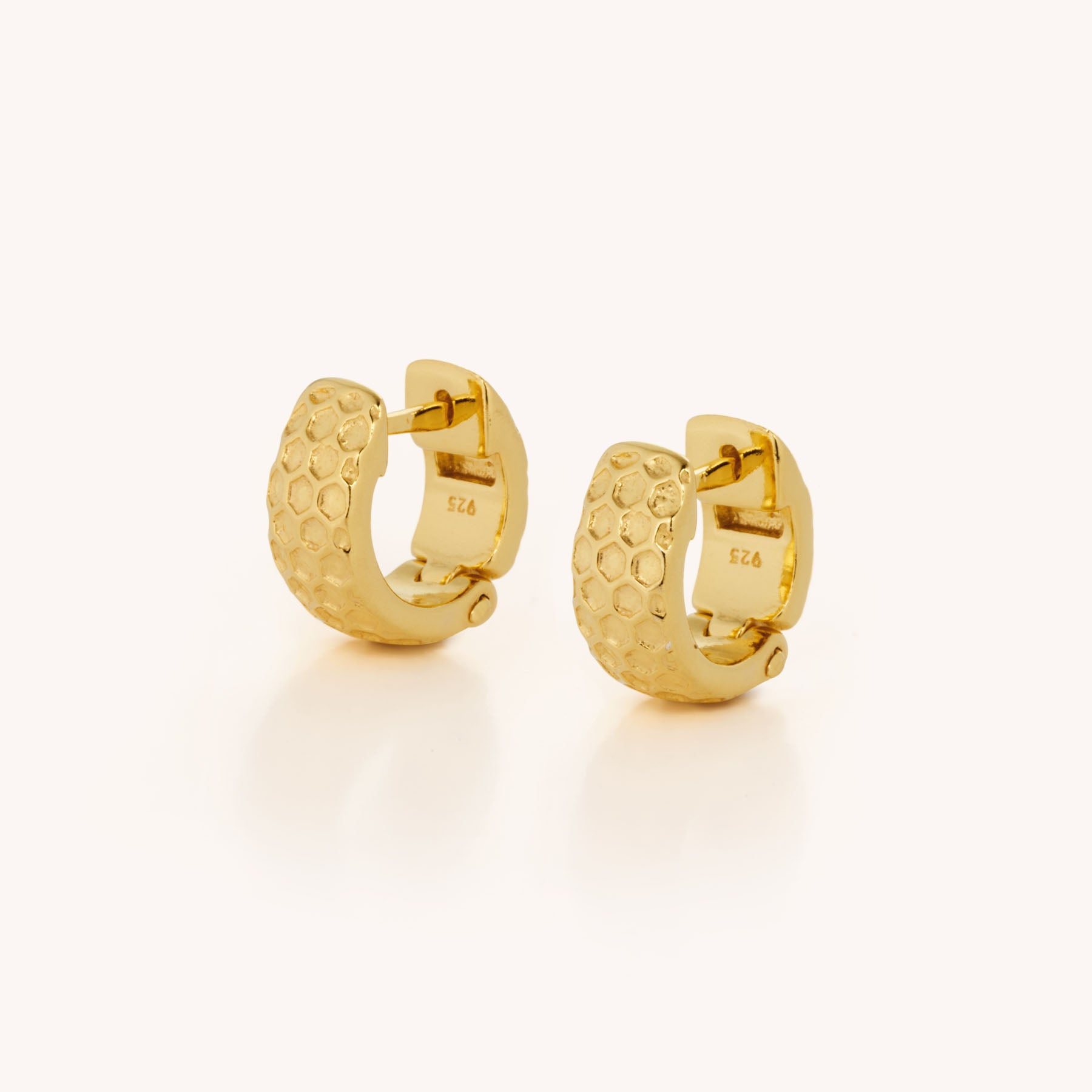 The Honeycomb Huggie Earrings are small gold hoops with a textured honeycomb pattern and hinged clasps, shown on a light background—ideal for anyone seeking stylish and durable earrings.