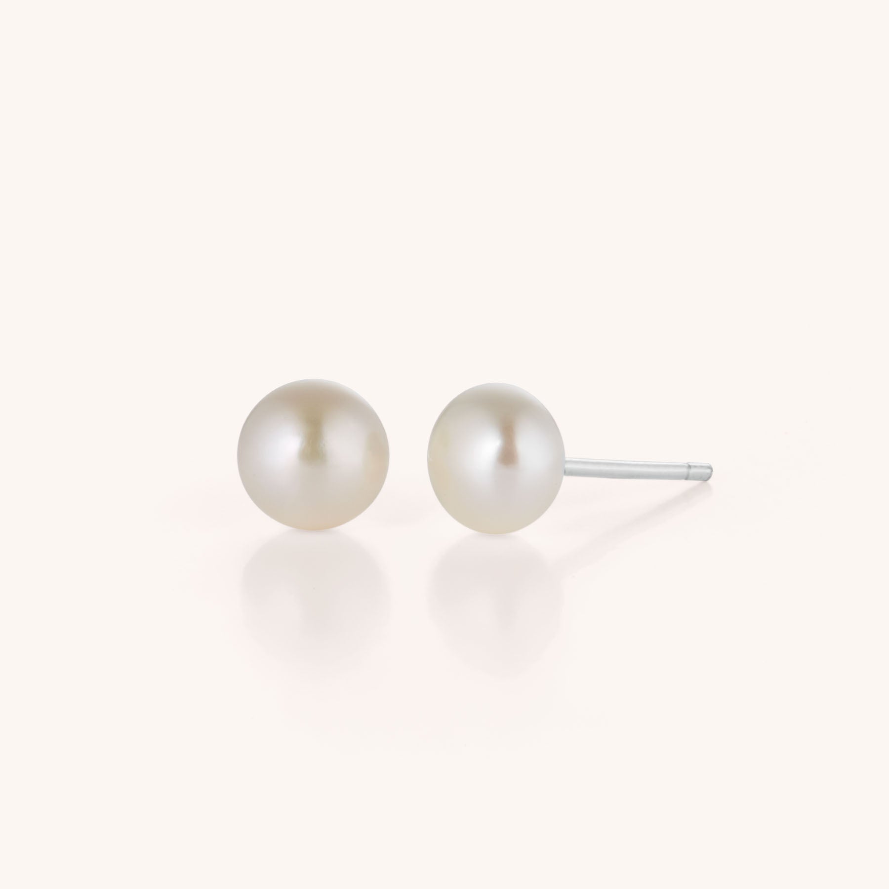 The Pearl Button Stud Earrings feature classic white pearls on silver posts, exuding timeless elegance and displayed against a simple light background.