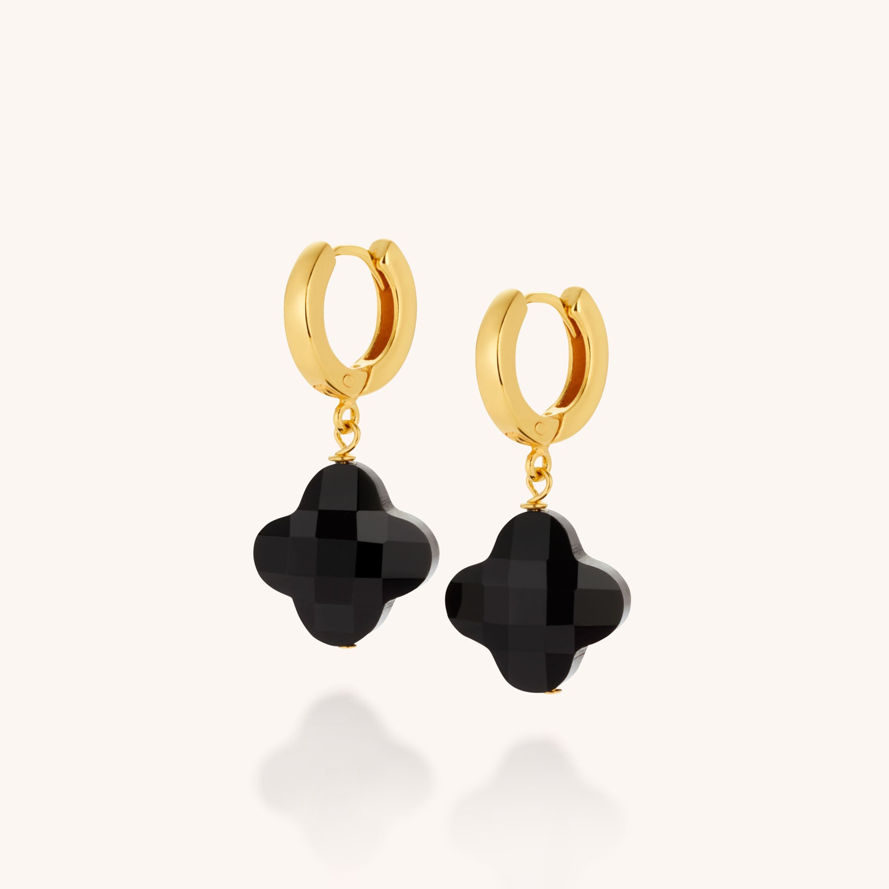 Clover Gem Hoop Earrings feature gold hoops with dangling black faceted quatrefoil gemstones, creating texture and shine. Displayed against a plain light background, these elegant earrings stand out with their unique clover design.