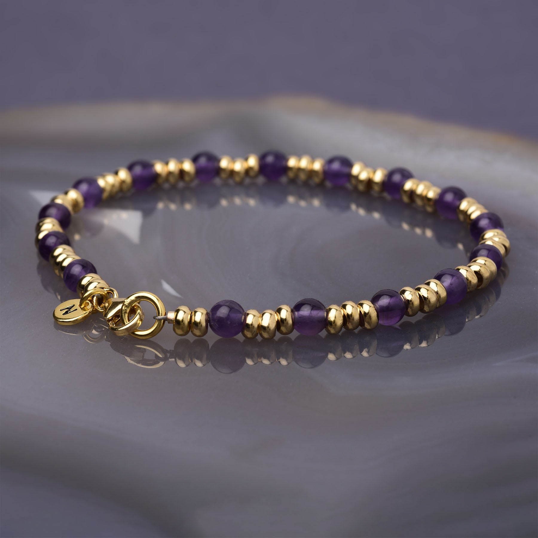 The Gem Bracelet February Birthstone features alternating gold and amethyst beads, a gold clasp, and a round charm—perfect for those who love February birthday or birthstone bracelets. Displayed on a marbled purple-gray surface.