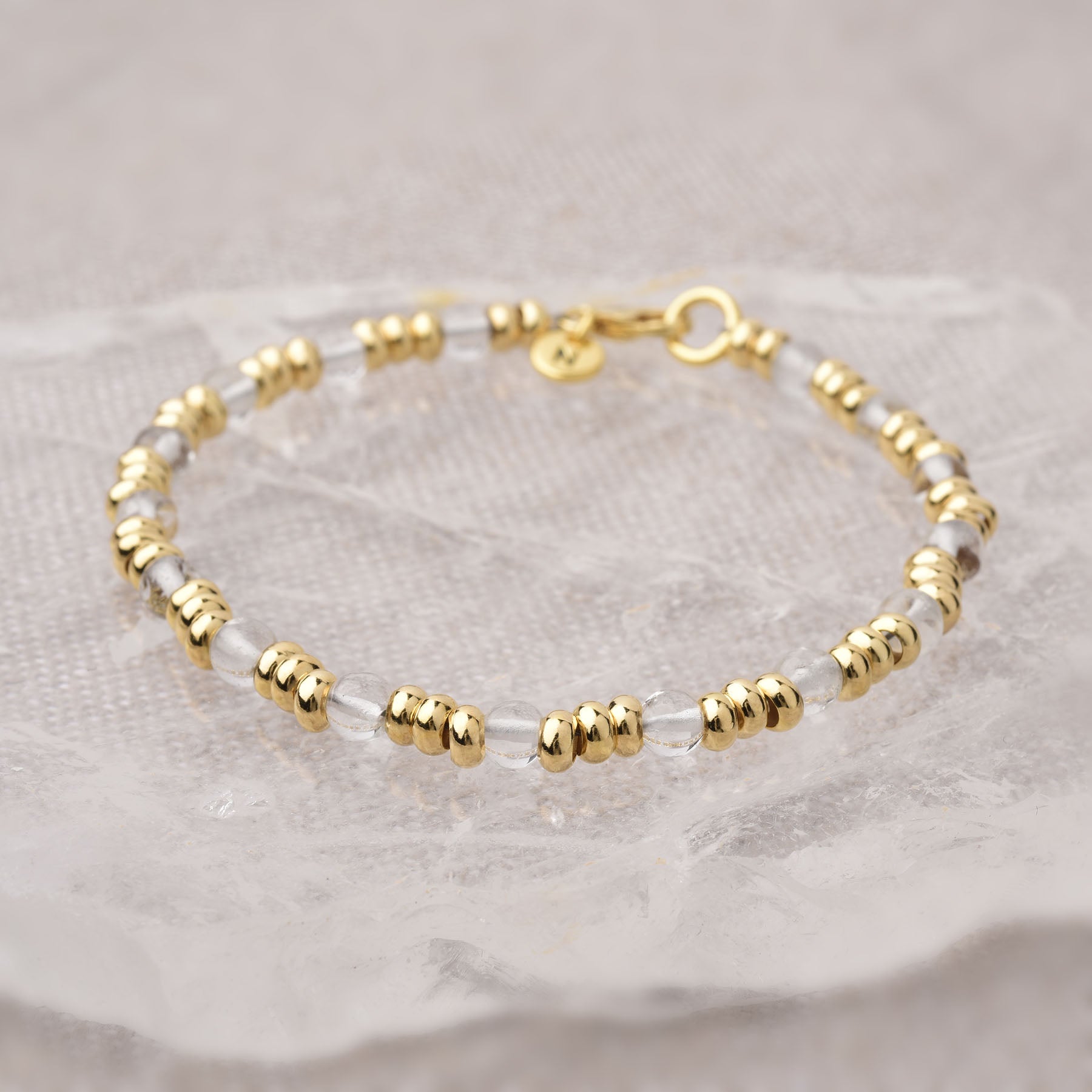 The Gem Bracelet March Birthstone features alternating small gold and clear beads, displayed on a textured surface, with a gold clasp and a tiny round gold charm near the closure.