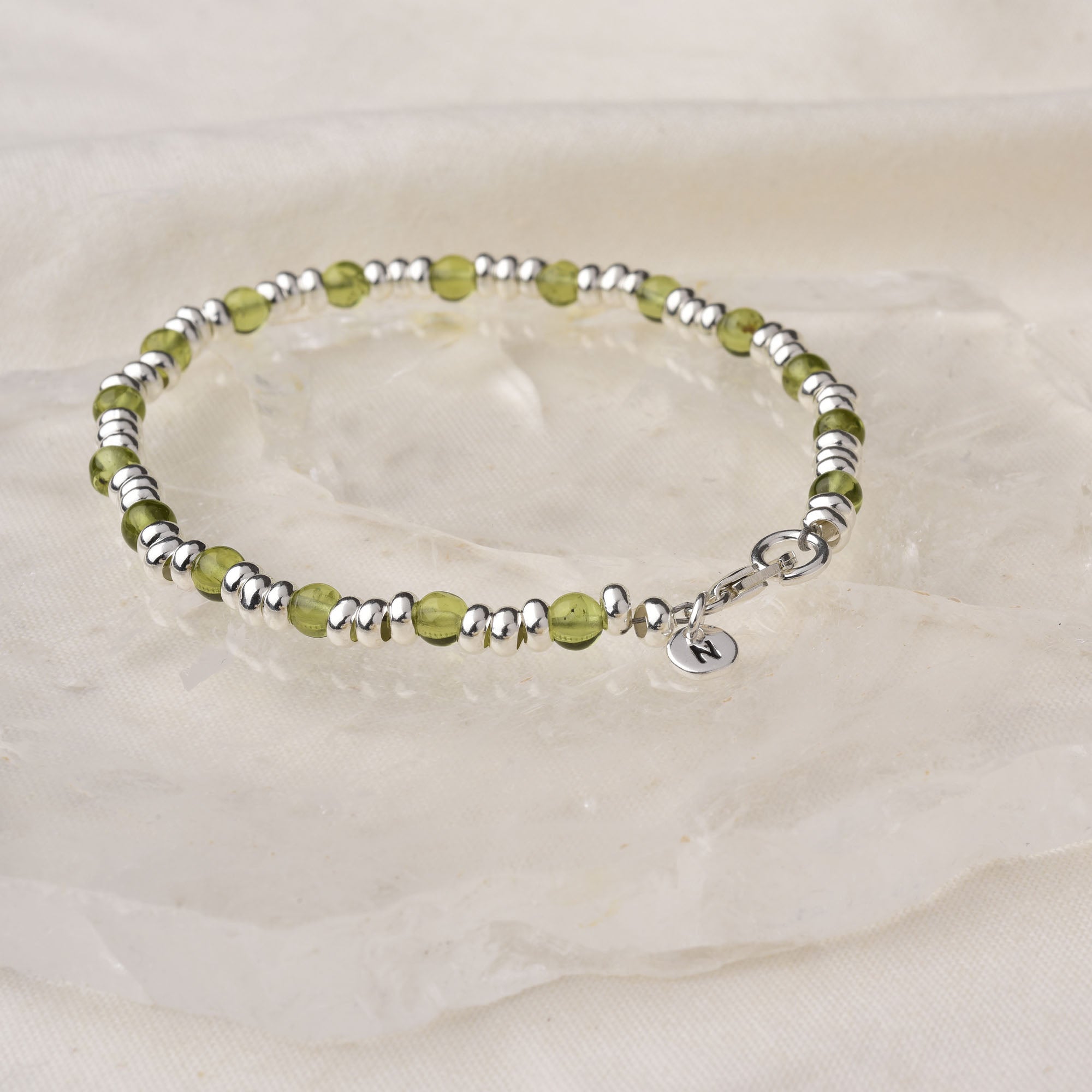 The Gem Birthstone Bracelet showcases alternating light green natural gemstone beads and eco sterling silver, finished with a round charm engraved with “N,” displayed on a clear crystal surface over cream-colored fabric.