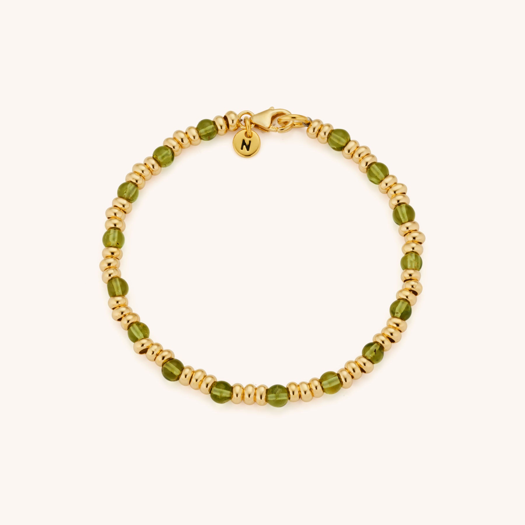 The Gem Birthstone Bracelet features alternating small gold and green natural gemstone beads, finished with a gold clasp and a round charm engraved with the letter N near the clasp.