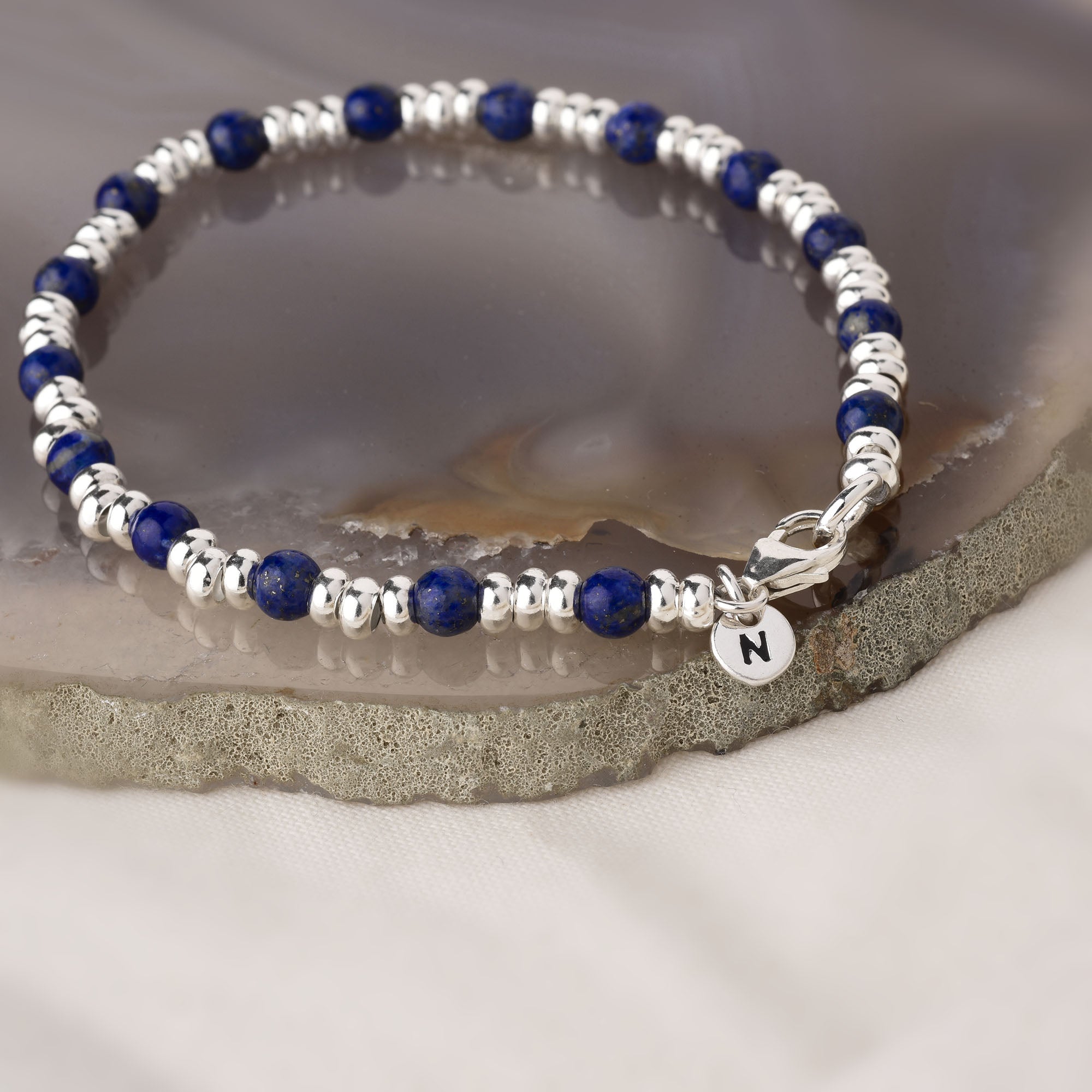 The Gem Bracelet September Birthstone features alternating silver and deep blue Lapis Lazuli beads, resting on a polished agate surface, with a small round silver charm engraved with the letter N near the clasp.