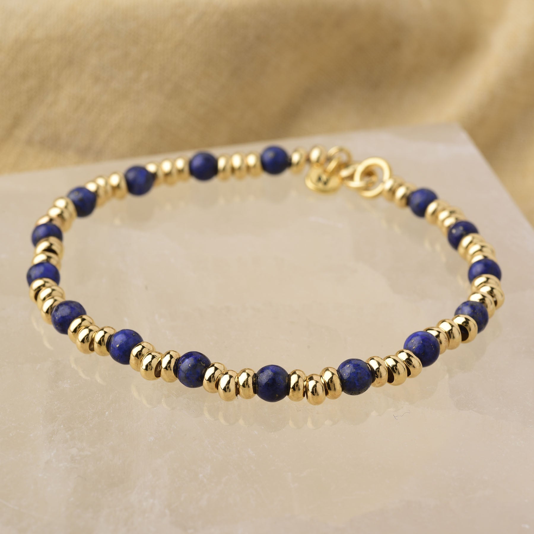 The Gem Bracelet September Birthstone features alternating round Lapis Lazuli gemstones and shiny gold beads, beautifully displayed on a reflective surface with a soft beige fabric background.