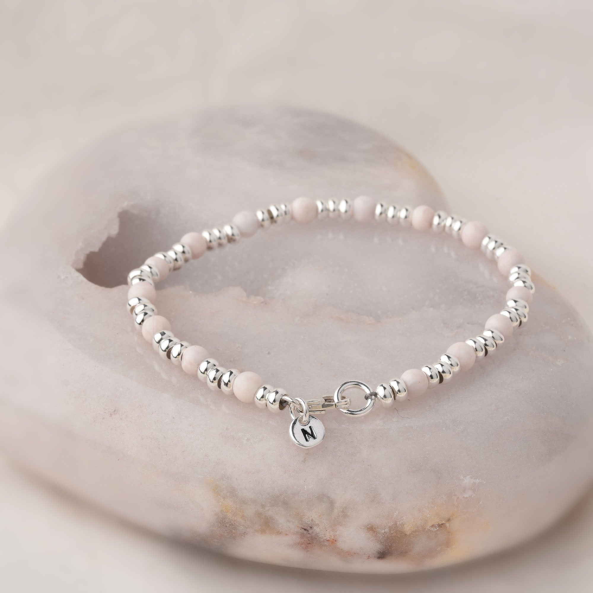 The Gem Bracelet October Birthstone features delicate pink opal beads and eco-silver spacers, accented with a round silver charm engraved with N, displayed on a smooth, light-colored stone.