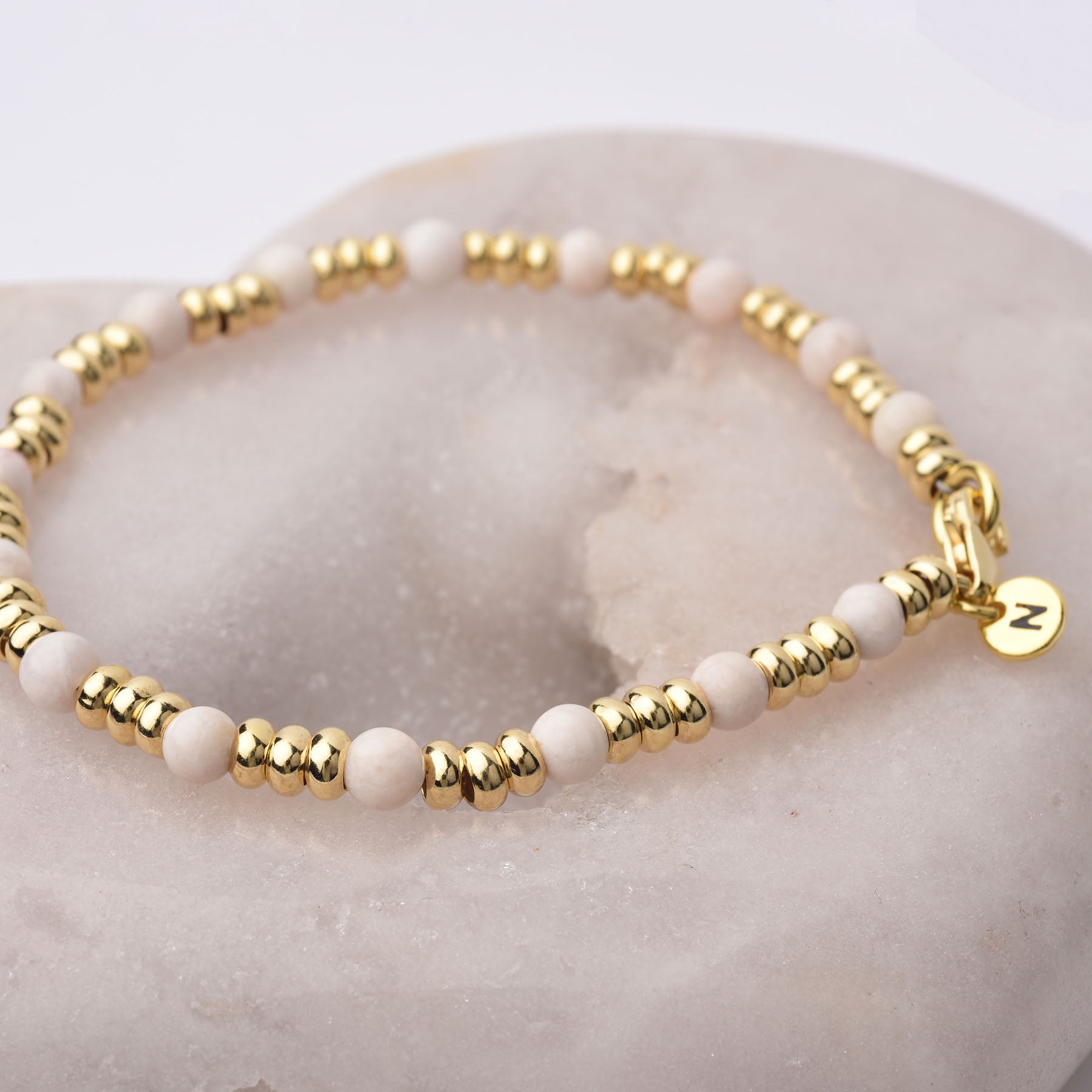 The Gem Bracelet October Birthstone features alternating small gold and Pink Opal beads, displayed on a smooth stone. A gold N charm hangs from the clasp, symbolizing emotional healing in this elegant accessory.
