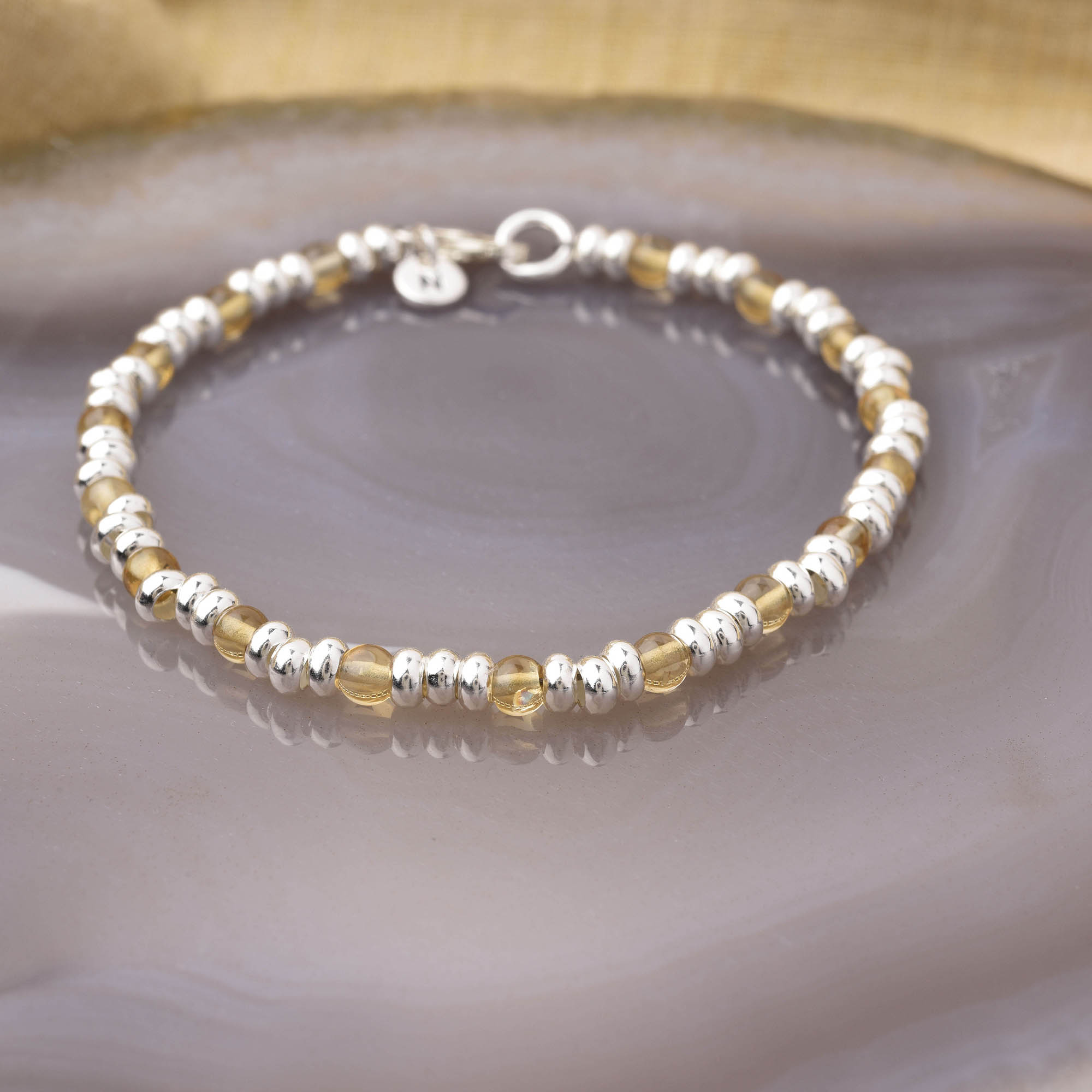The Gem Bracelet November Birthstone features alternating small silver and citrine gemstone beads, displayed on a polished surface with a gold-toned background—ideal for adding stylish abundance to your look.