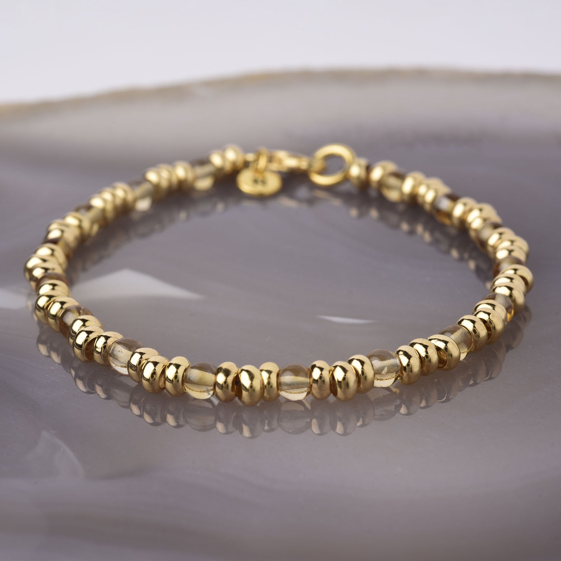 The Gem Bracelet November Birthstone, a gold beaded bracelet with shiny metallic, transparent beads, and Citrine gemstone accents for abundance, is shown on a glossy surface against a soft, neutral background.
