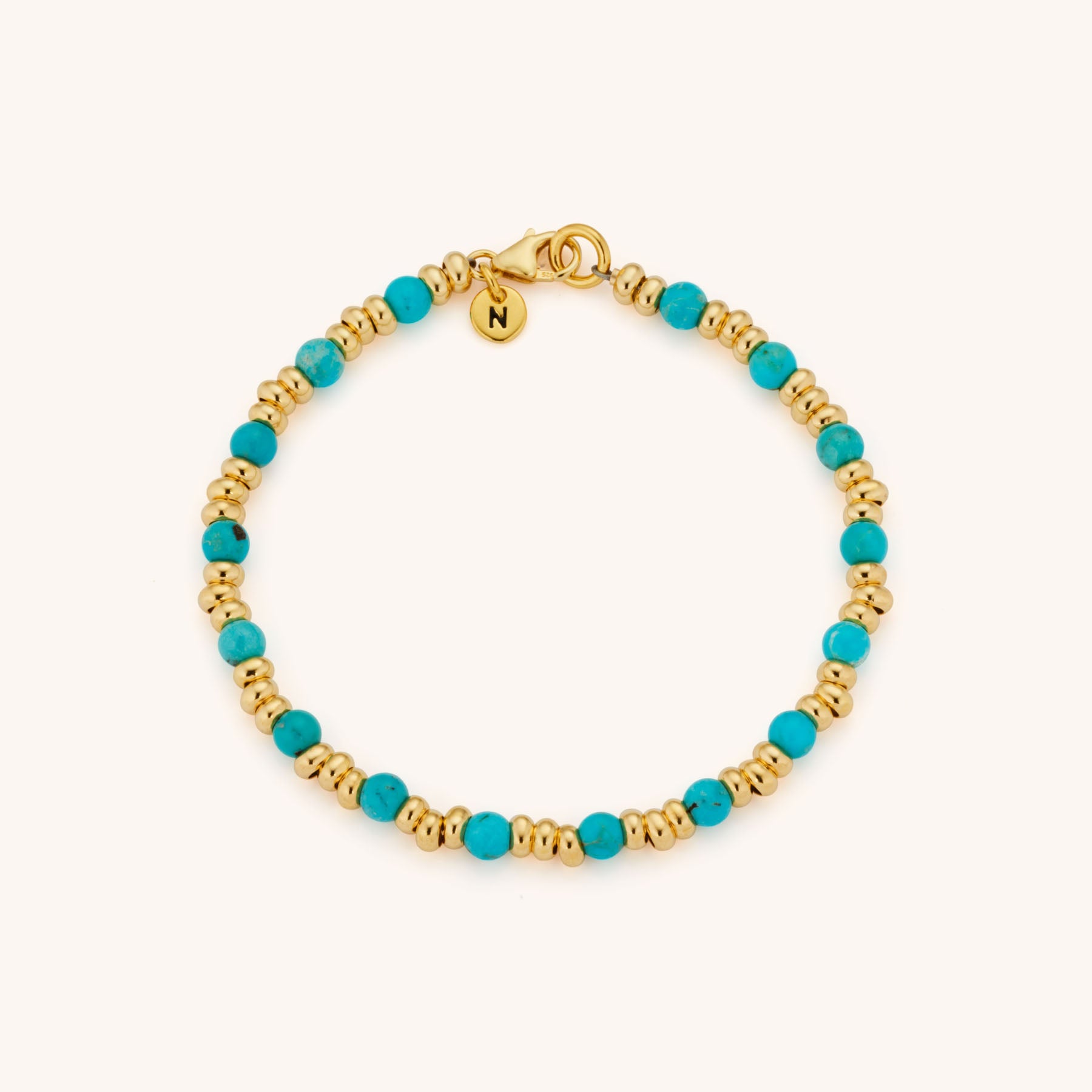 The Gem Bracelet December Birthstone features gold and turquoise beads with a small round gold charm engraved with N. Turquoise, the December birthstone, adds vibrant color to this elegant accessory. Displayed on a plain light background.