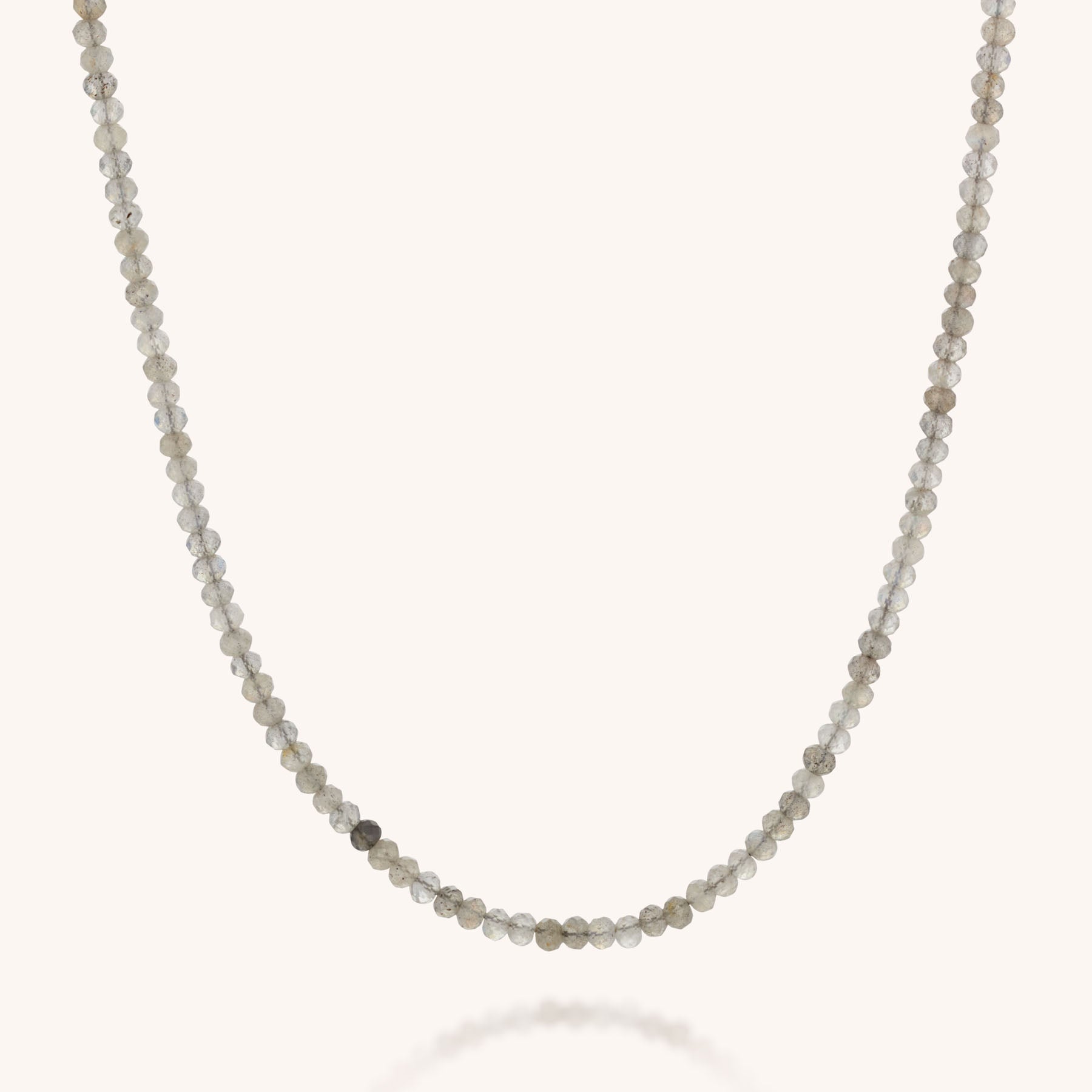 The Mini Bead Necklace features small, round faceted beads in light gray and beige on a single strand, finished with a sterling silver clasp, set against a plain light background.
