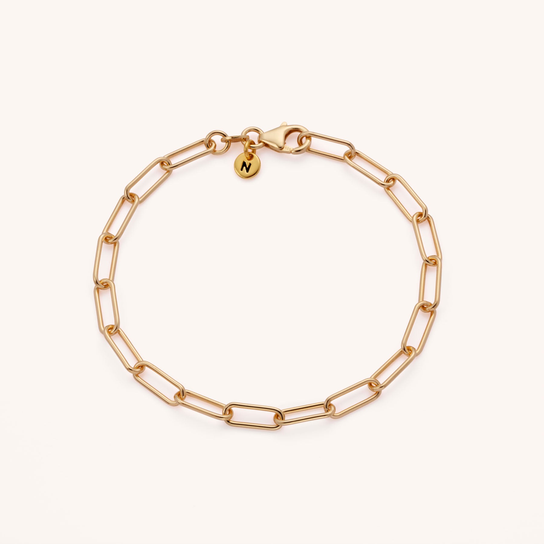 The Paperclip Bracelet features delicate elongated rectangular links, a clasp, and a small round charm engraved with the letter N. Its adjustable length design is shown on a plain, light background.