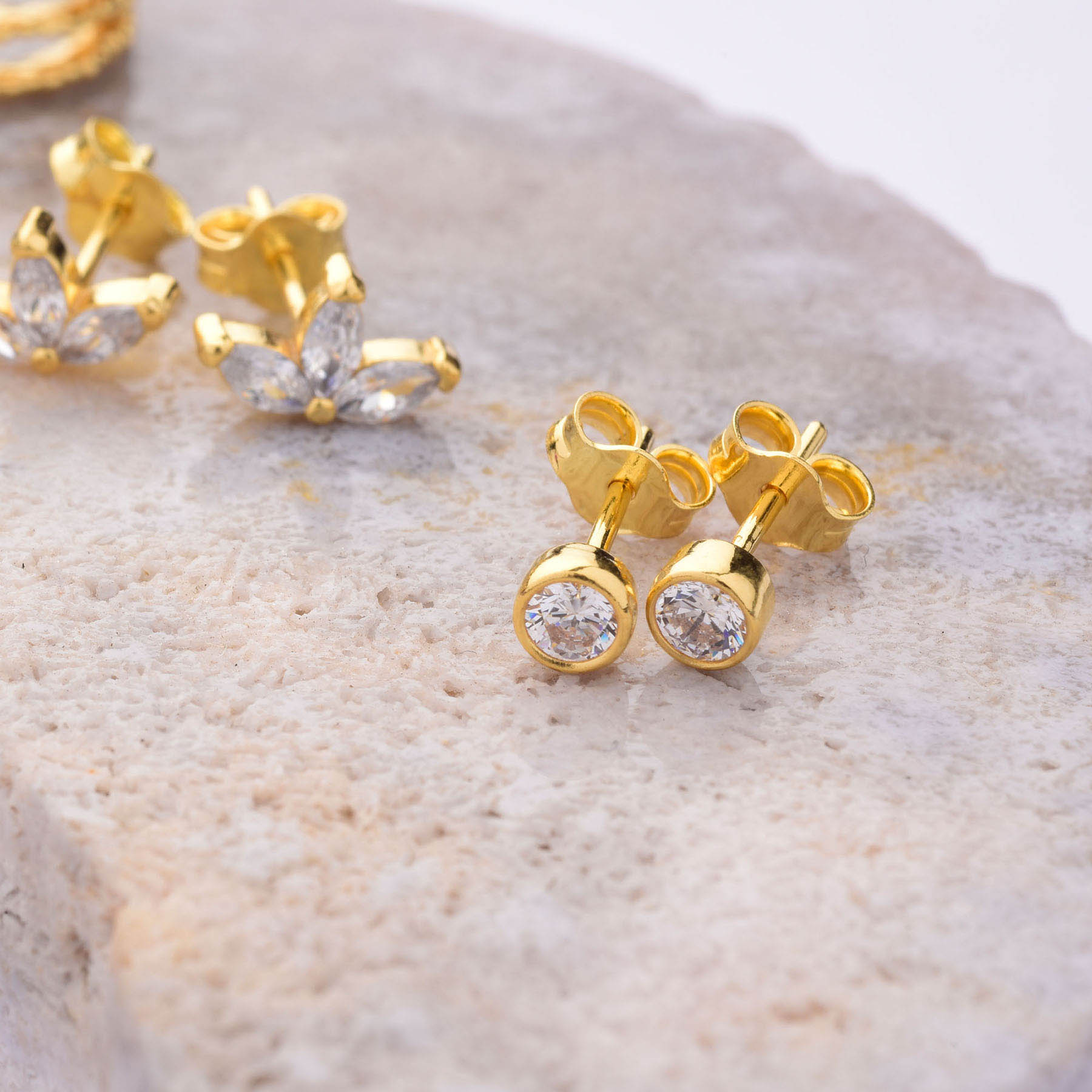 Dot Stud Earrings in gold with round clear stones are shown on a textured stone surface, while a second pair of gold earrings with marquise-shaped stones appears in the background.