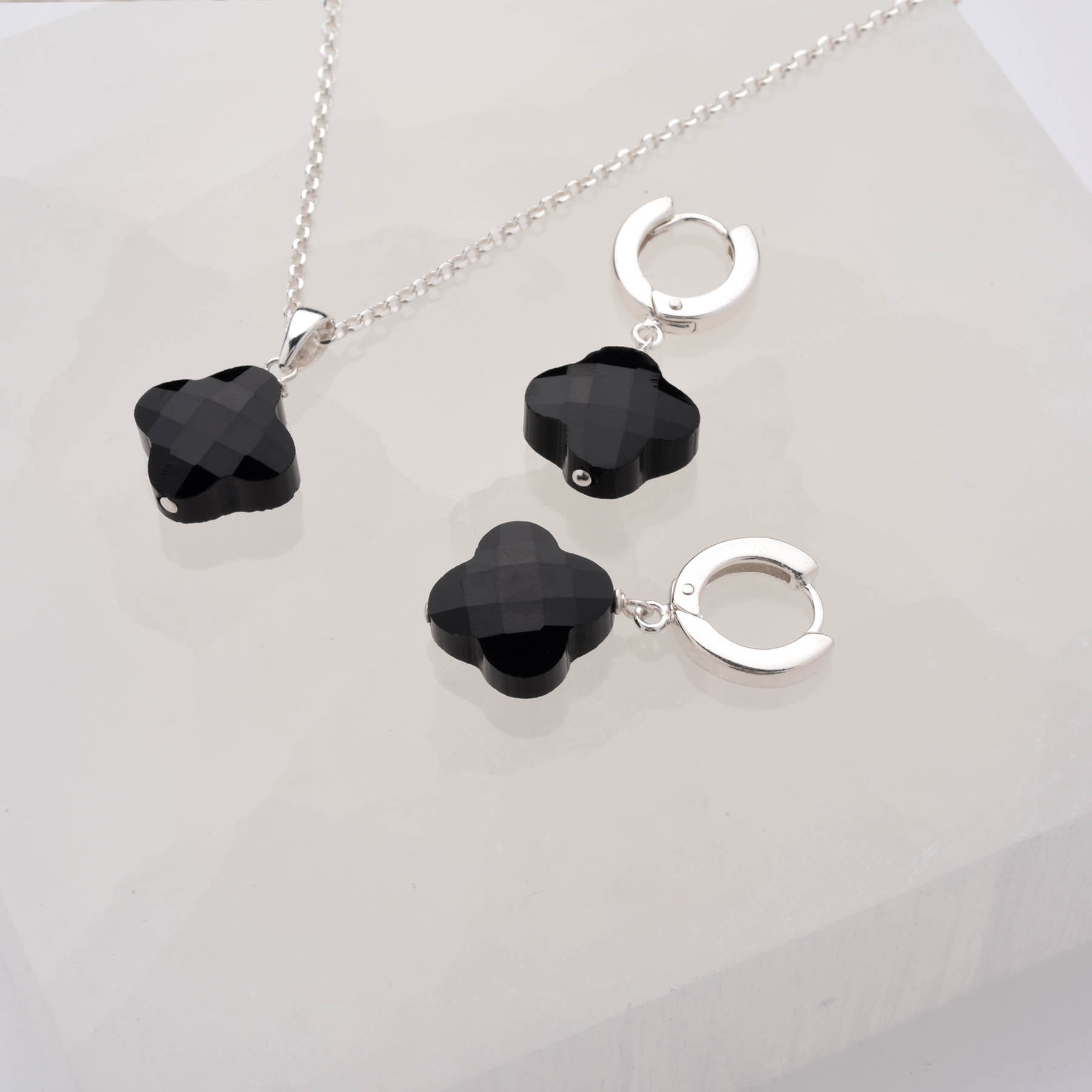 The Clover Gem Hoop Earrings in sterling silver, featuring faceted black gemstones, are displayed with a matching necklace on a white geometric surface.