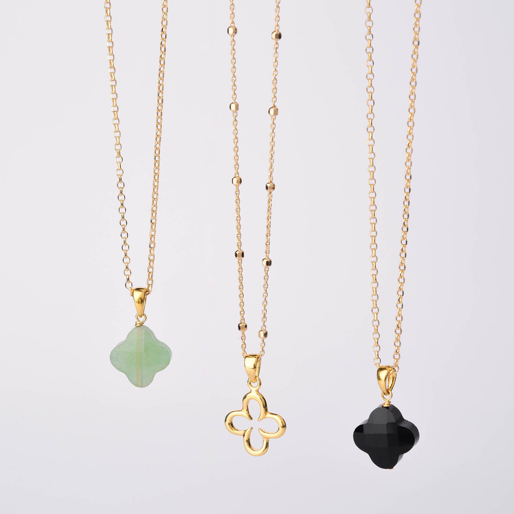 Three Gem Clover Necklaces hang vertically on a white background, each with a clover pendant: one green gemstone, one gold outline, and one black. The center necklaces chain features small gold bead accents.