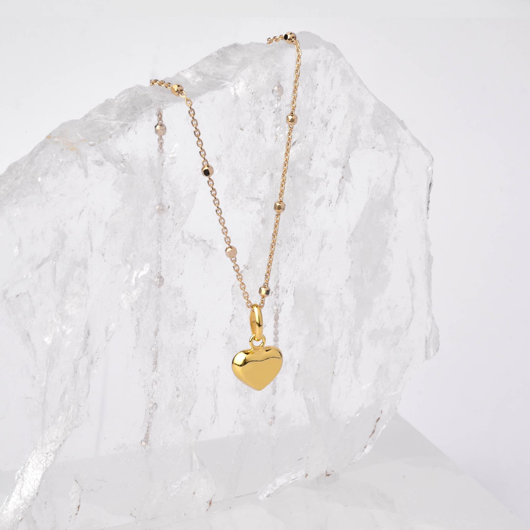 The Heart Charm Necklace, featuring a gold plated chain and a small heart pendant, is displayed draped over a large textured crystal or glass piece on a plain white background.