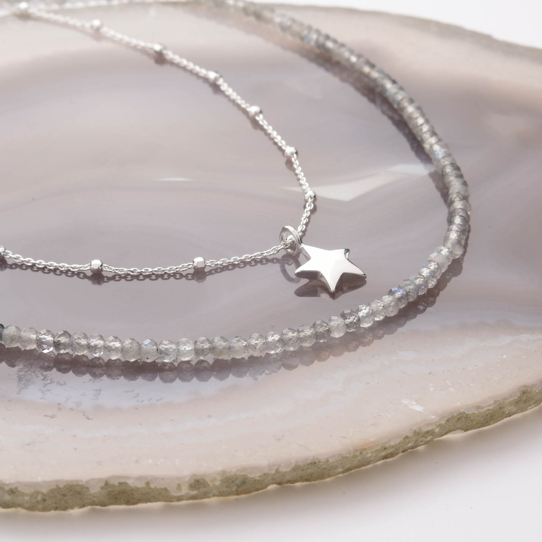 Two Mini Bead Necklaces rest on a polished agate slice: one features faceted gray beads with a sterling silver clasp, and the other is a silver chain adorned with a small star pendant.