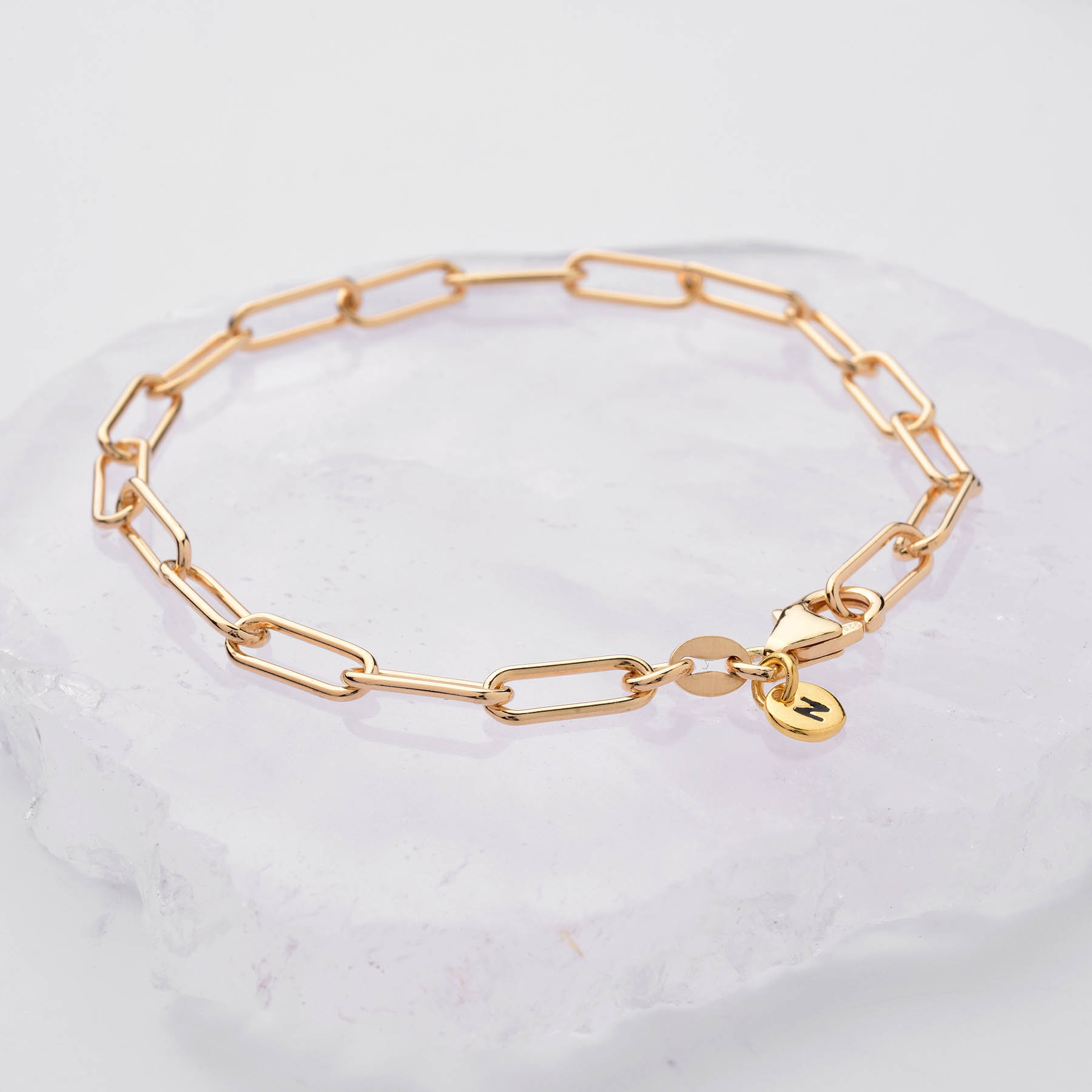 The Paperclip Bracelet features a gold, elongated link chain and a small round charm engraved with M. Displayed on a clear, textured surface, this bracelet offers an adjustable length for the perfect fit.