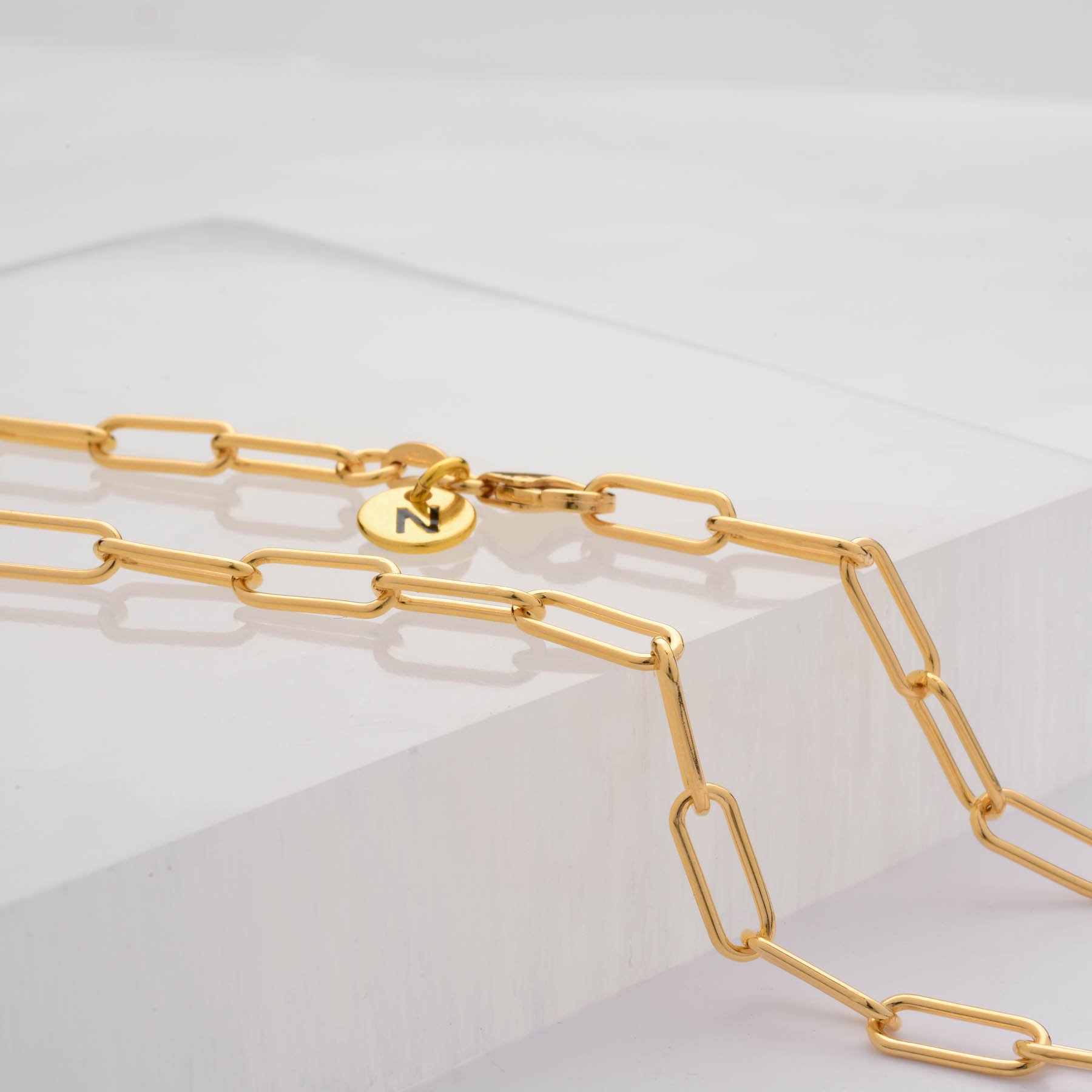 The Paperclip Necklace features elongated rectangular gold links and a round N-stamped charm, displayed on clear acrylic—a modern piece ideal for those who love elevated style.