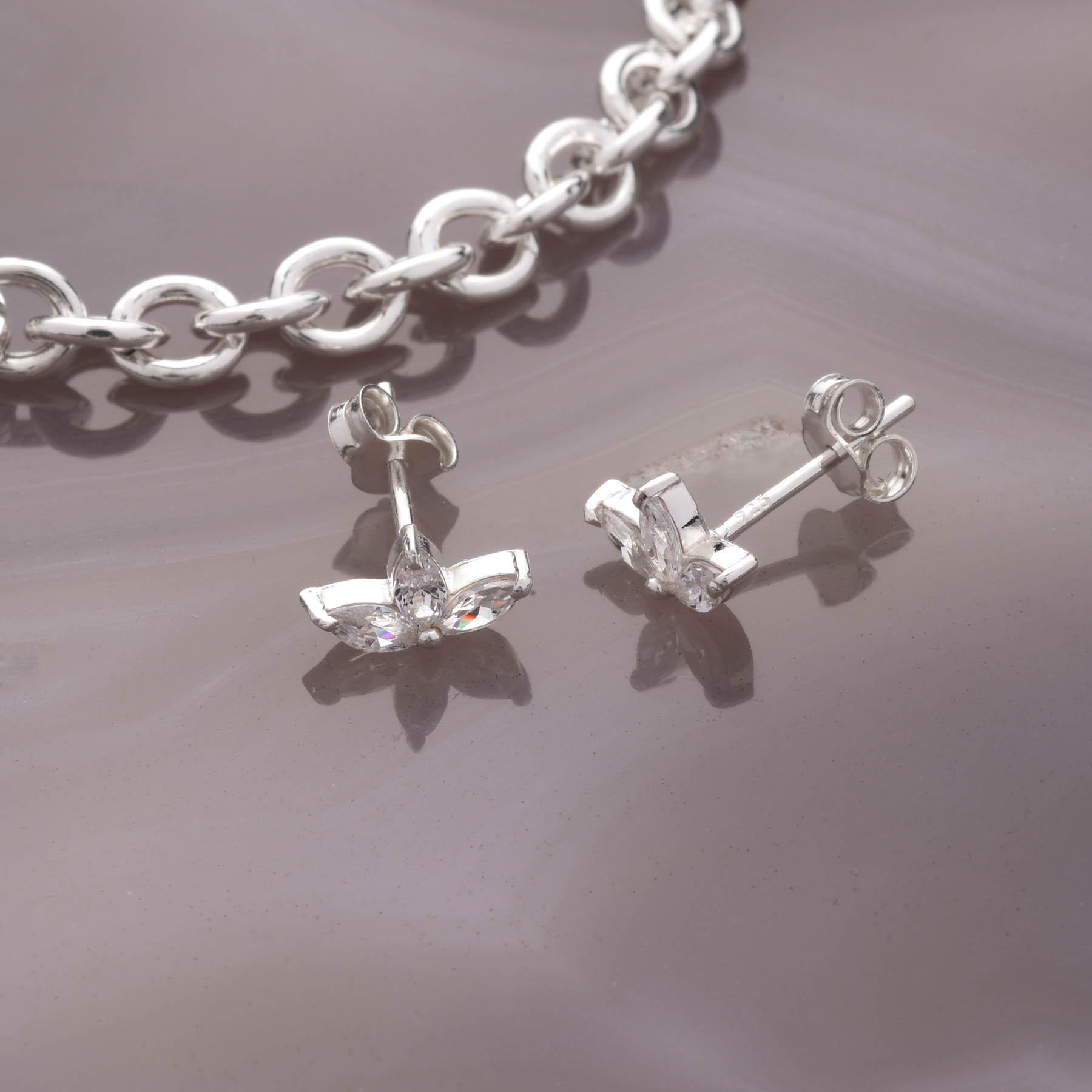 The Trio Leaf Stud Earrings, crafted from 925 silver and set with clear cubic zirconia, are displayed on a smooth marbled surface beside a chunky silver chain bracelet.