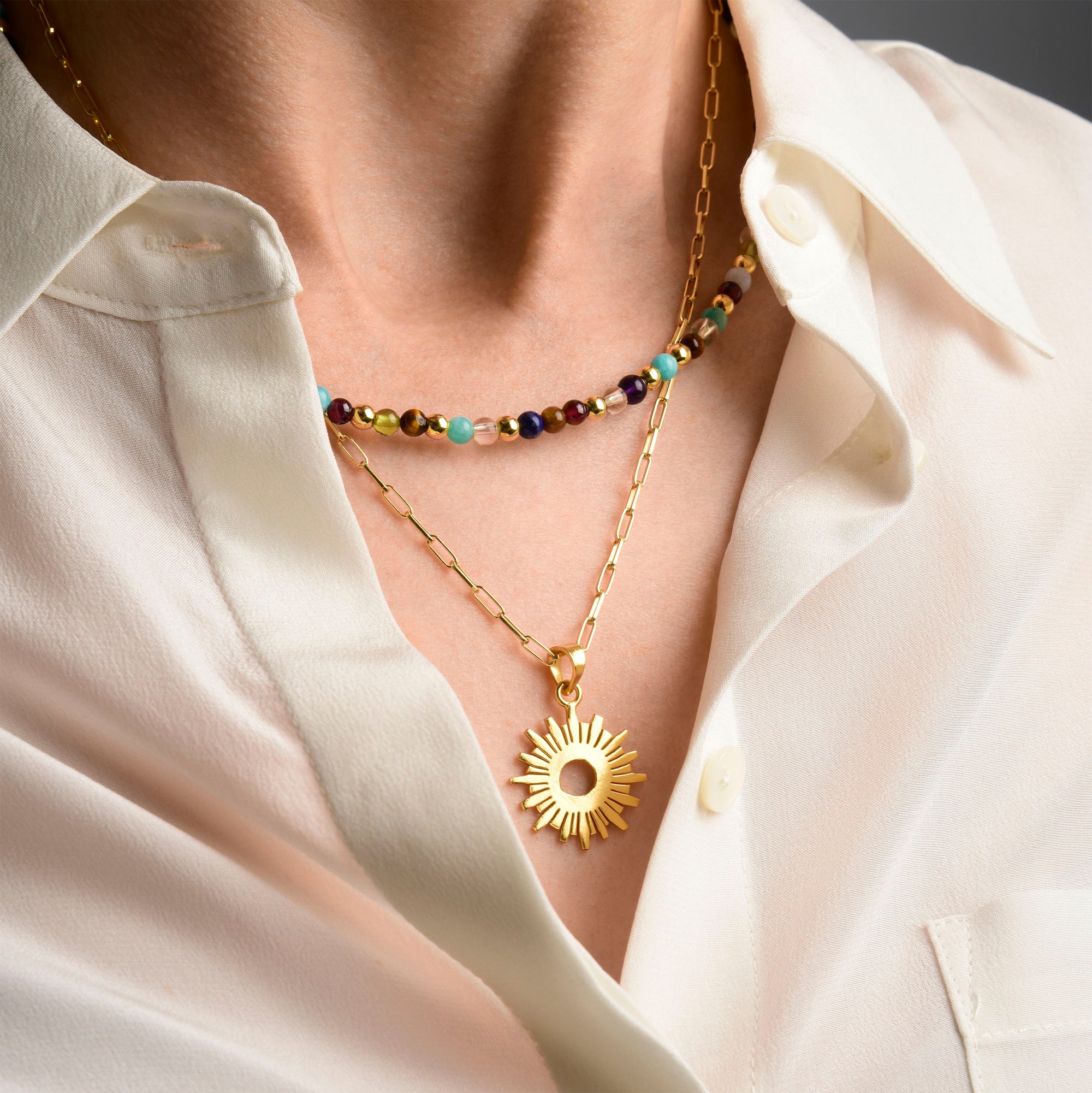 A person wearing a cream button-up shirt with two layered necklaces: one with colorful beads and another gold chain with a sun-shaped pendant.