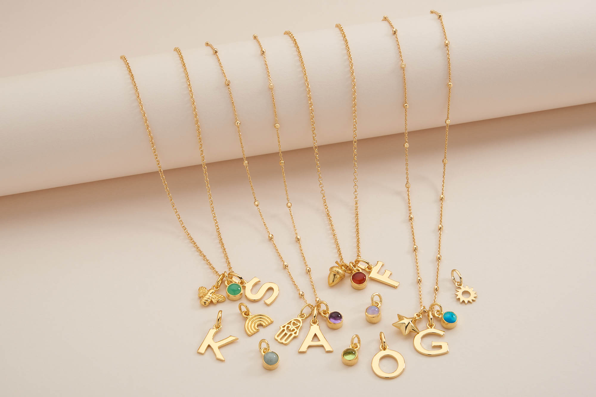 Five gold necklaces with large letter pendants (K, S, A, O, G), each adorned with colorful charms and beads, displayed on a cream-colored background.