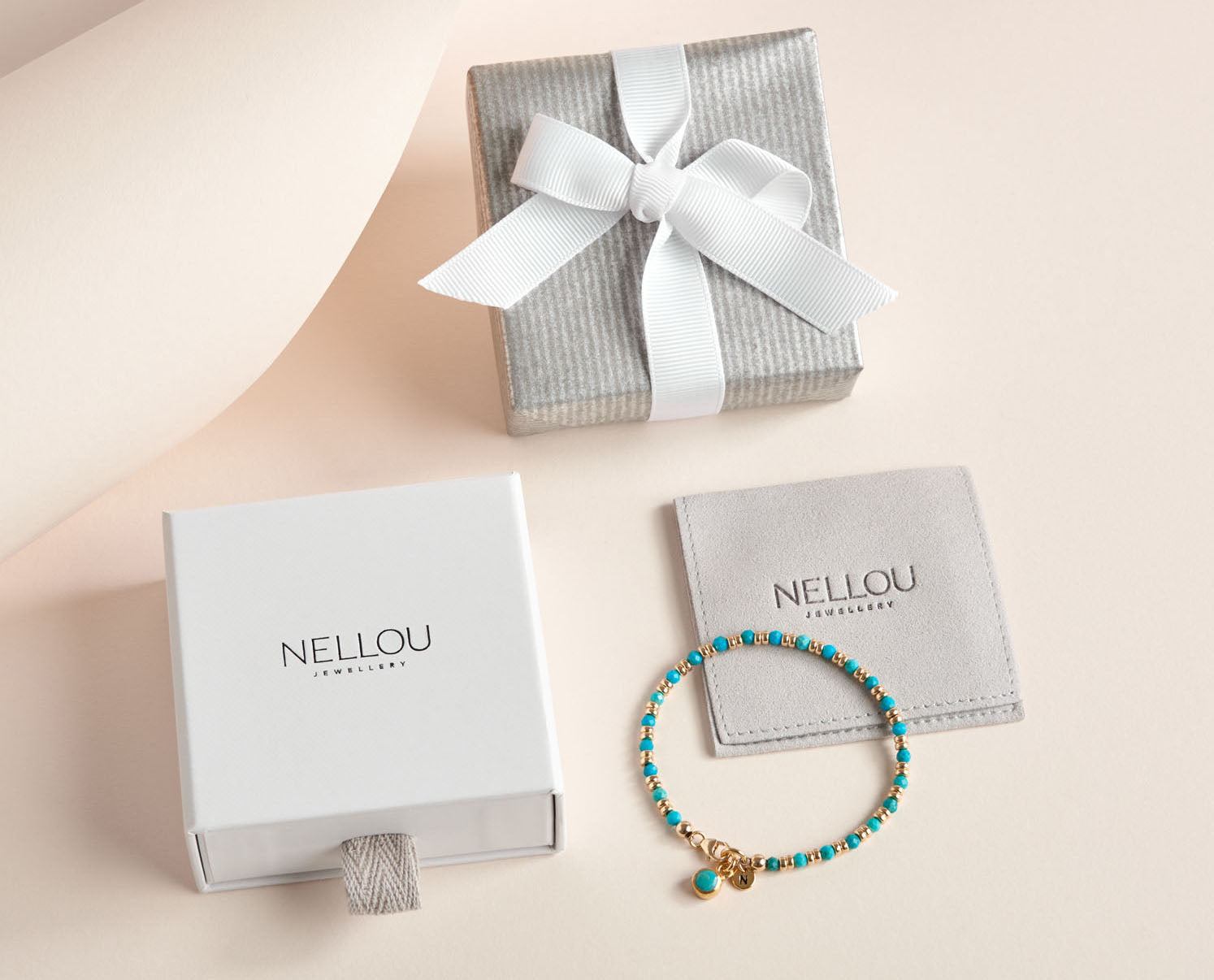 A turquoise and gold beaded bracelet with a charm is displayed next to a gray gift box, a white NELLOU Jewelry box, and a branded gray pouch on a light beige background.