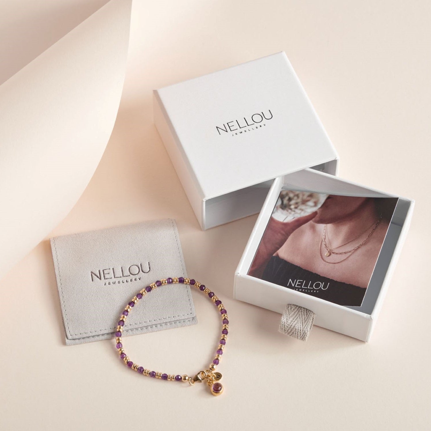A gold and purple beaded bracelet with a charm, displayed next to a grey pouch and an open white NELLOU Jewellery box with a jewelry photo inside, all on a pale background.