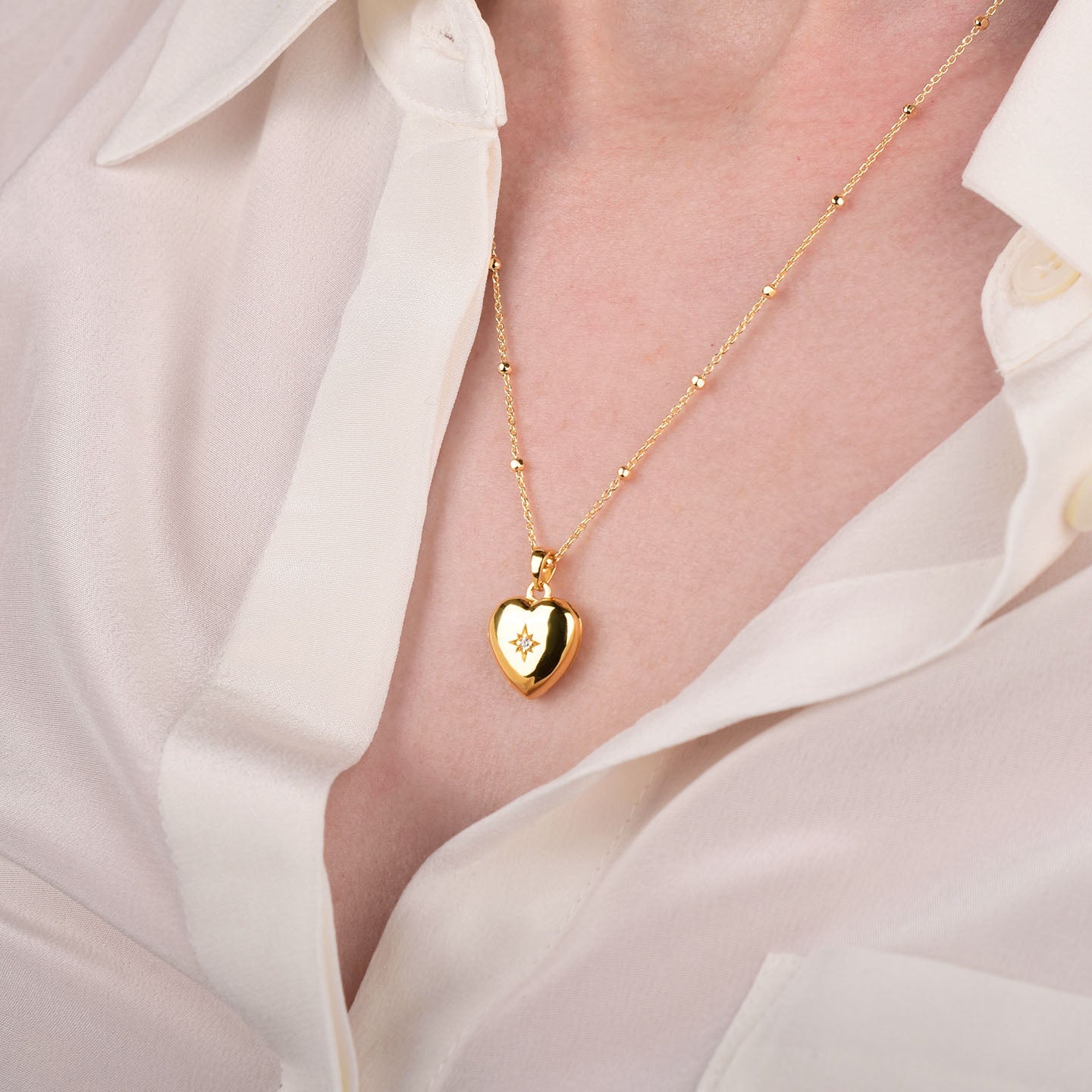 A person wearing a cream-colored blouse with a gold heart-shaped pendant necklace featuring a small star design, shown close-up on their chest.