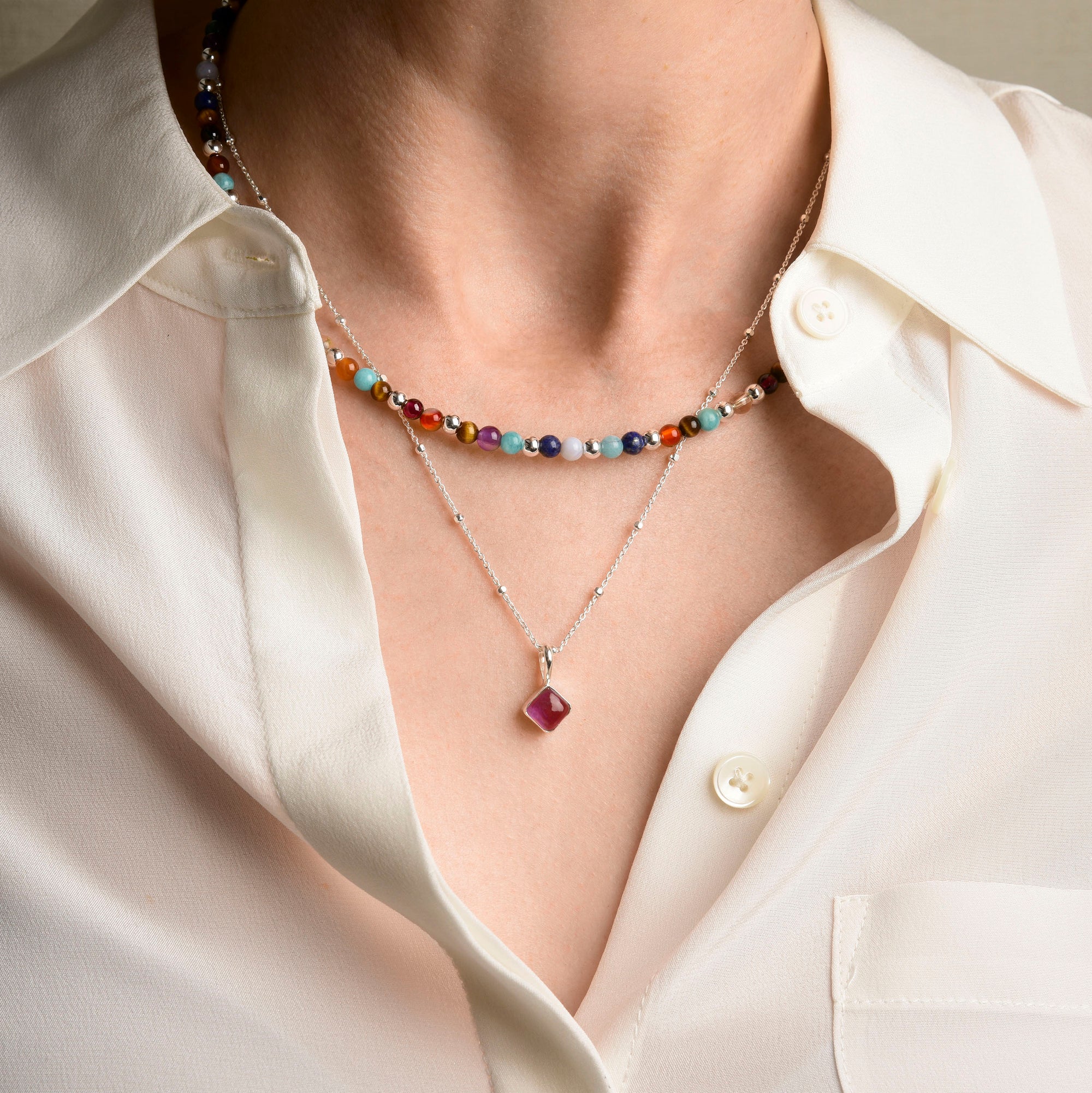 A person wearing a white button-up shirt and two necklaces: one with colorful beads and the other with a silver chain and a purple stone pendant.