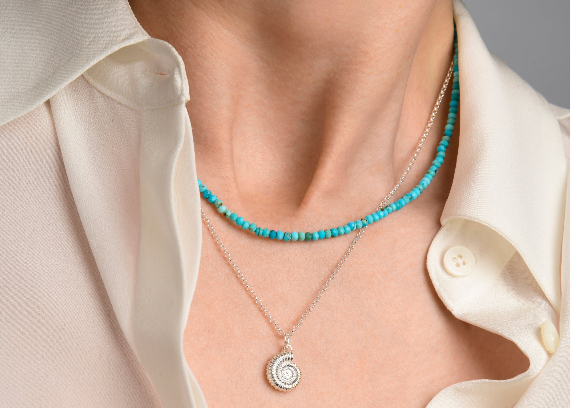 Close-up of a person wearing a white button-up shirt, a turquoise beaded necklace, and a silver chain necklace with a spiral pendant.
