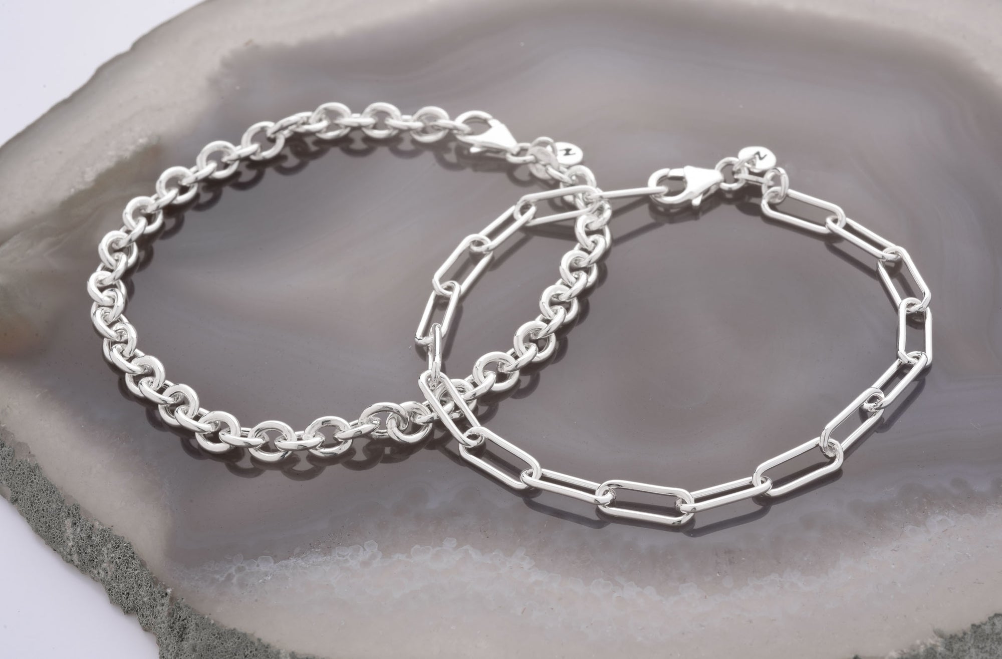 Two silver chain bracelets, one with round links and the other with elongated oval links, displayed on a polished gray agate stone surface.
