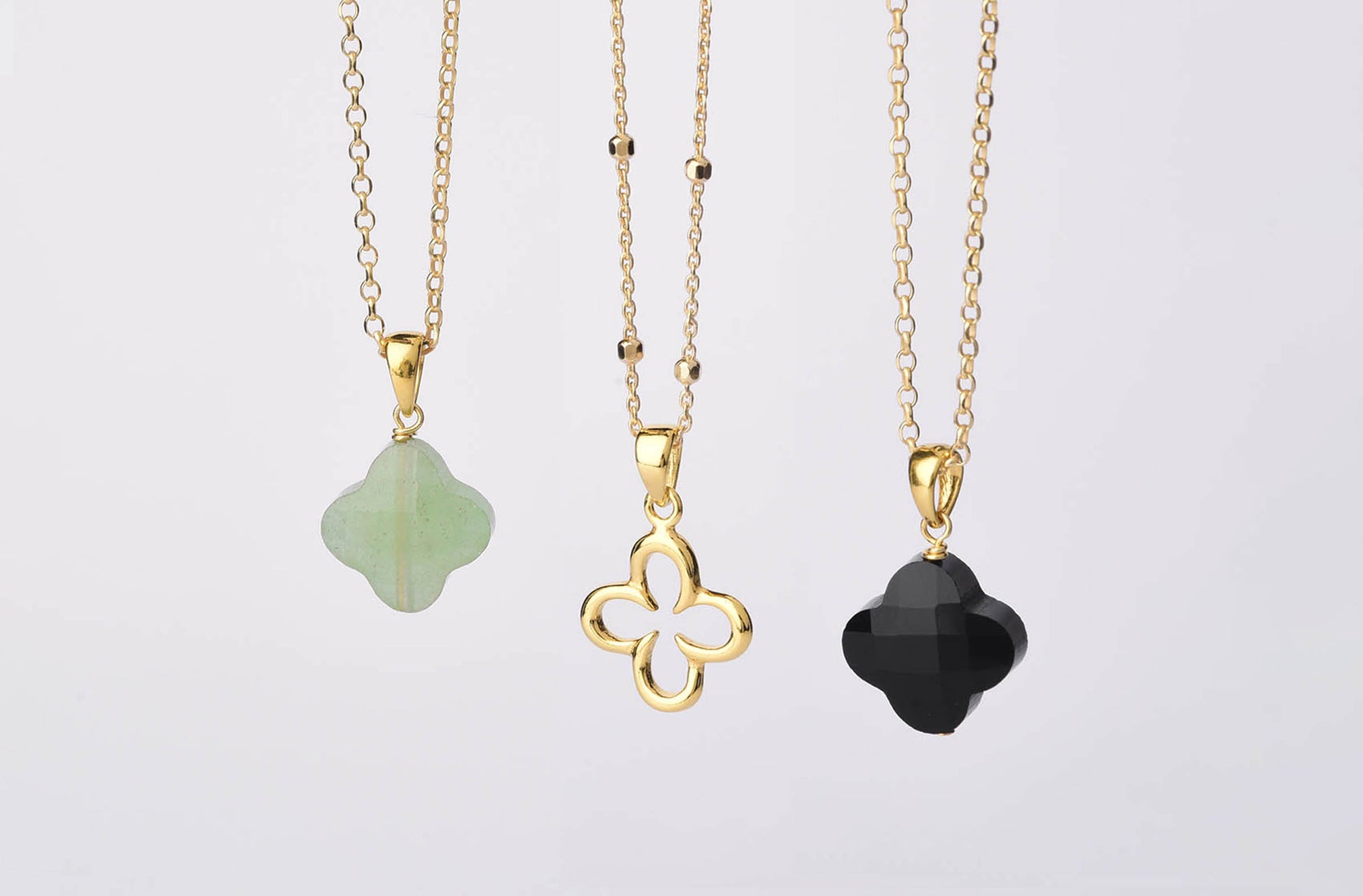 Three gold necklaces with pendants: a green clover-shaped stone, a gold outline clover, and a black faceted clover-shaped stone, all hanging against a plain white background.