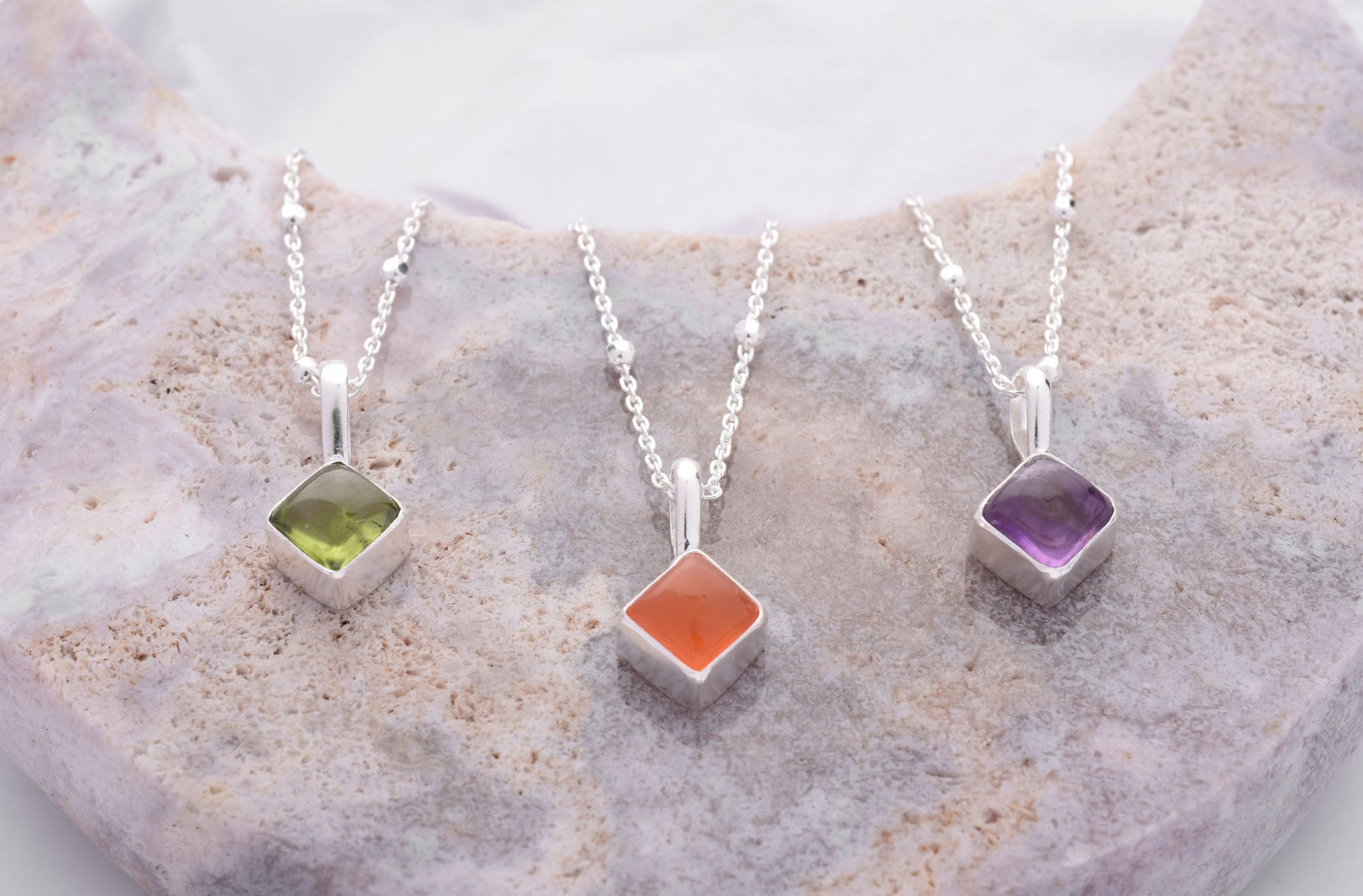 Three silver necklaces with square gemstone pendants—green, orange, and purple—are displayed on a pale, textured stone background.